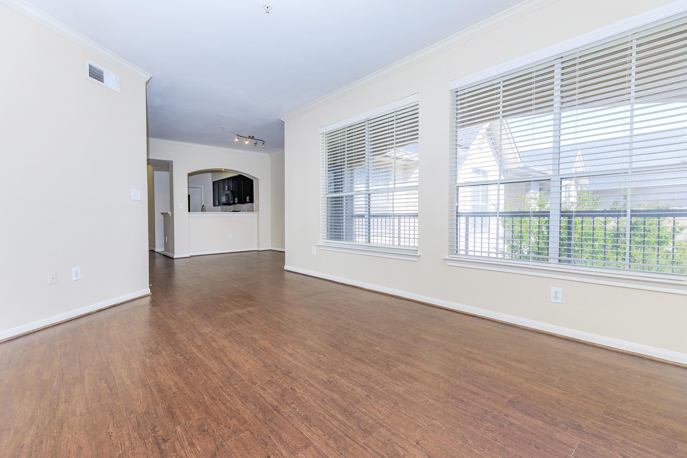Spacious, empty room with light-colored walls and wood-style flooring. Large windows with blinds allow natural light to fill the space. An archway leads to a kitchen area in the background, creating an inviting, open layout. Ideal for decorating or furnishing to suit personal style.