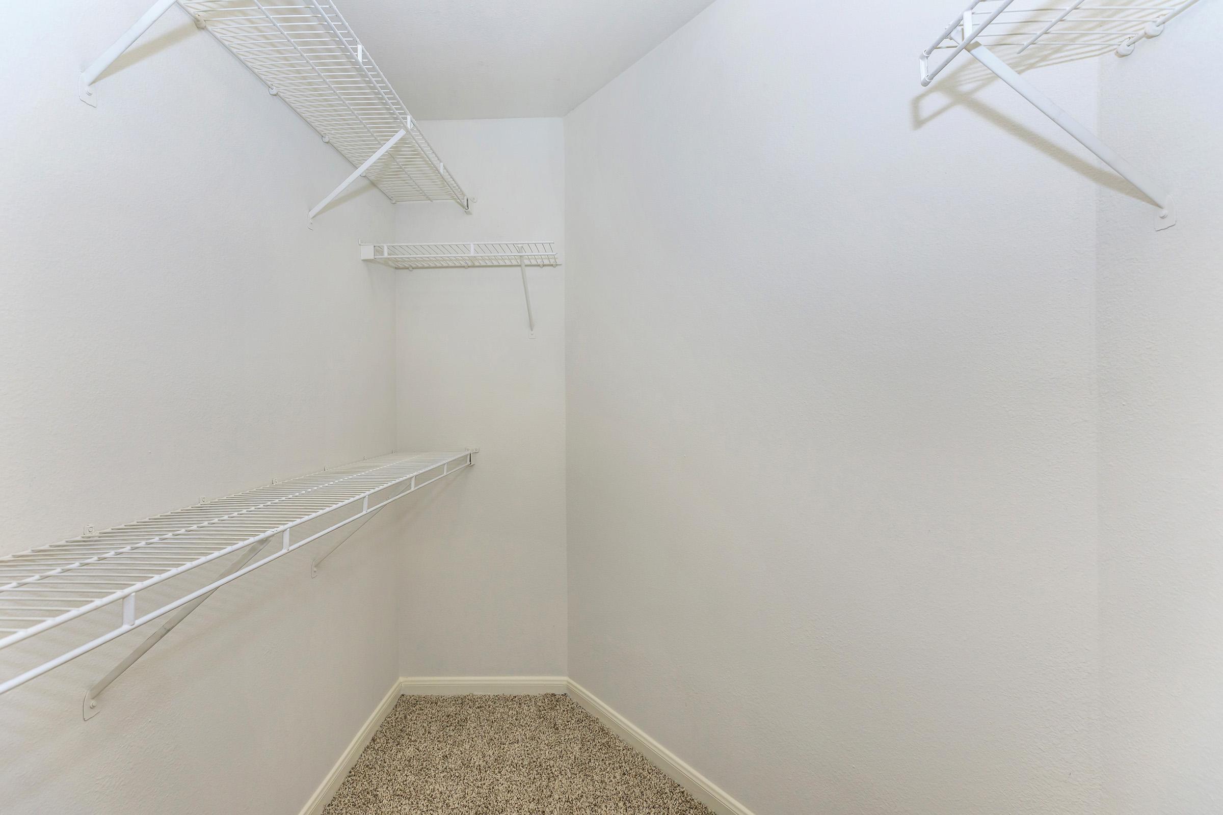 Empty closet with white wire shelving on the walls. The walls are plain and white, and the floor is covered with a light-colored carpet. The closet has a small and minimalist design, with no items stored inside.