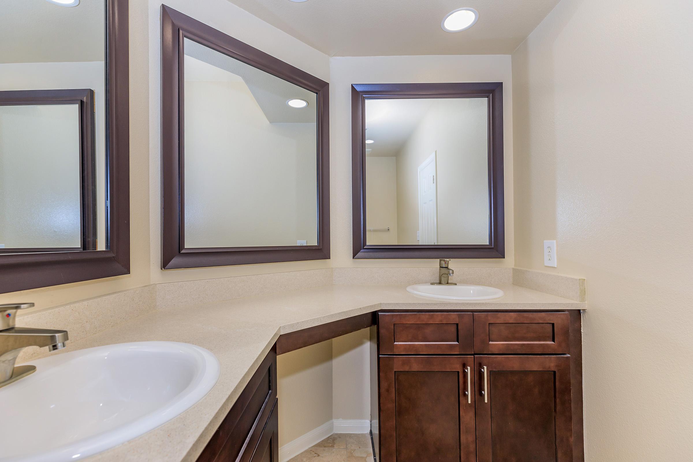 A bright, modern bathroom featuring double sinks with chrome faucets, dark wooden cabinetry, and two large square mirrors above each sink. The walls are painted a light beige, and the flooring is a light-colored tile. The overall design is clean and contemporary.