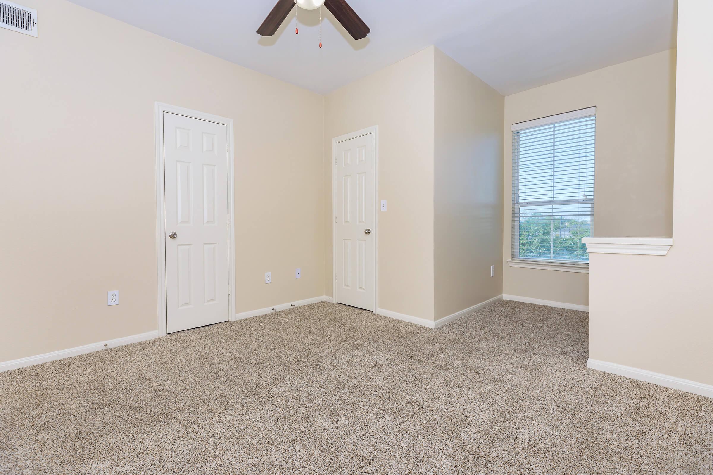 Empty room featuring light beige walls, two closed doors on opposite walls, a ceiling fan with wood blades, and a window with blinds allowing natural light in. The flooring is covered with light brown carpet.