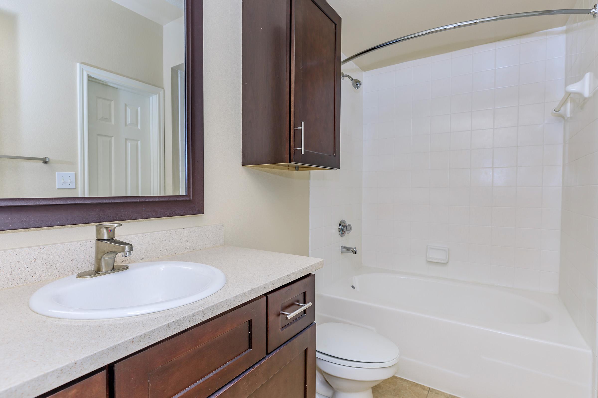 A clean and modern bathroom featuring a white bathtub with a shower curtain, a vanity with a sink, a mirror, and dark wood cabinets. The walls are a light beige, and there's a towel bar, providing a bright and tidy appearance.