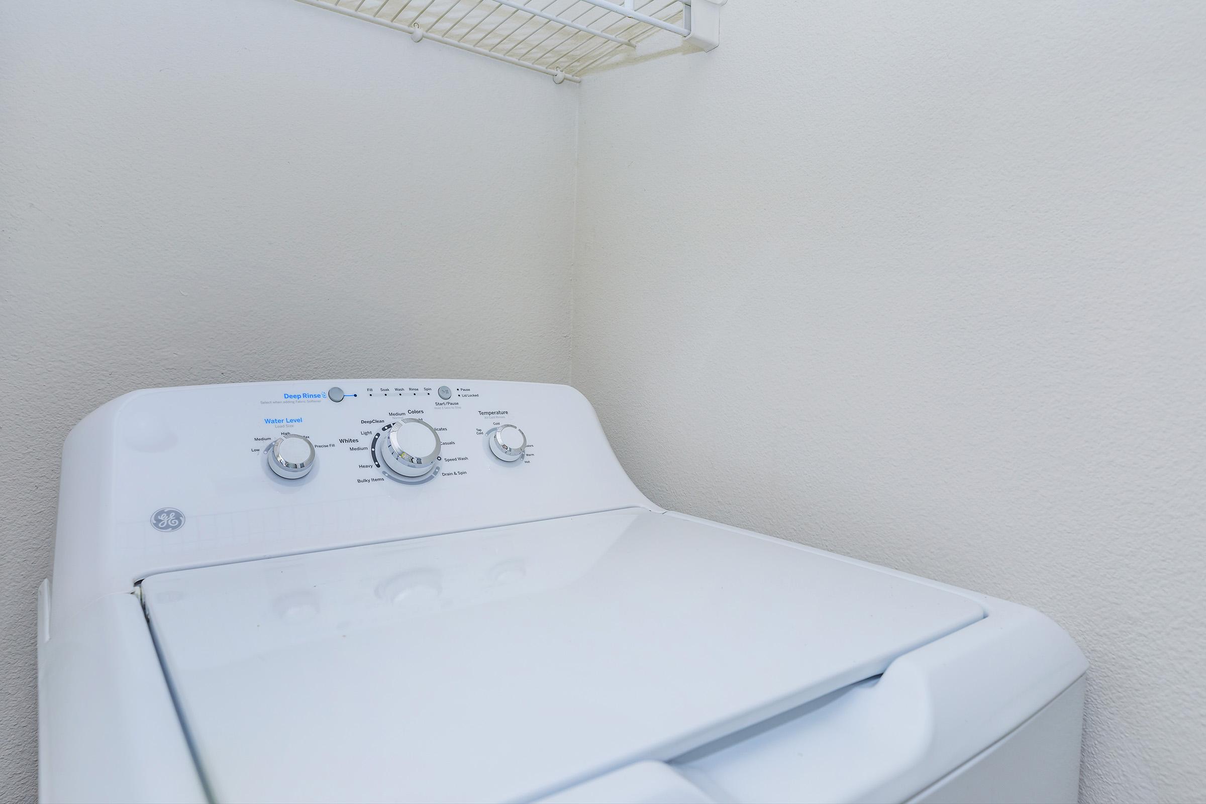 A top-loading washing machine with control dials and a lid open, situated in a corner against a plain light-colored wall, with a wire shelf above it.