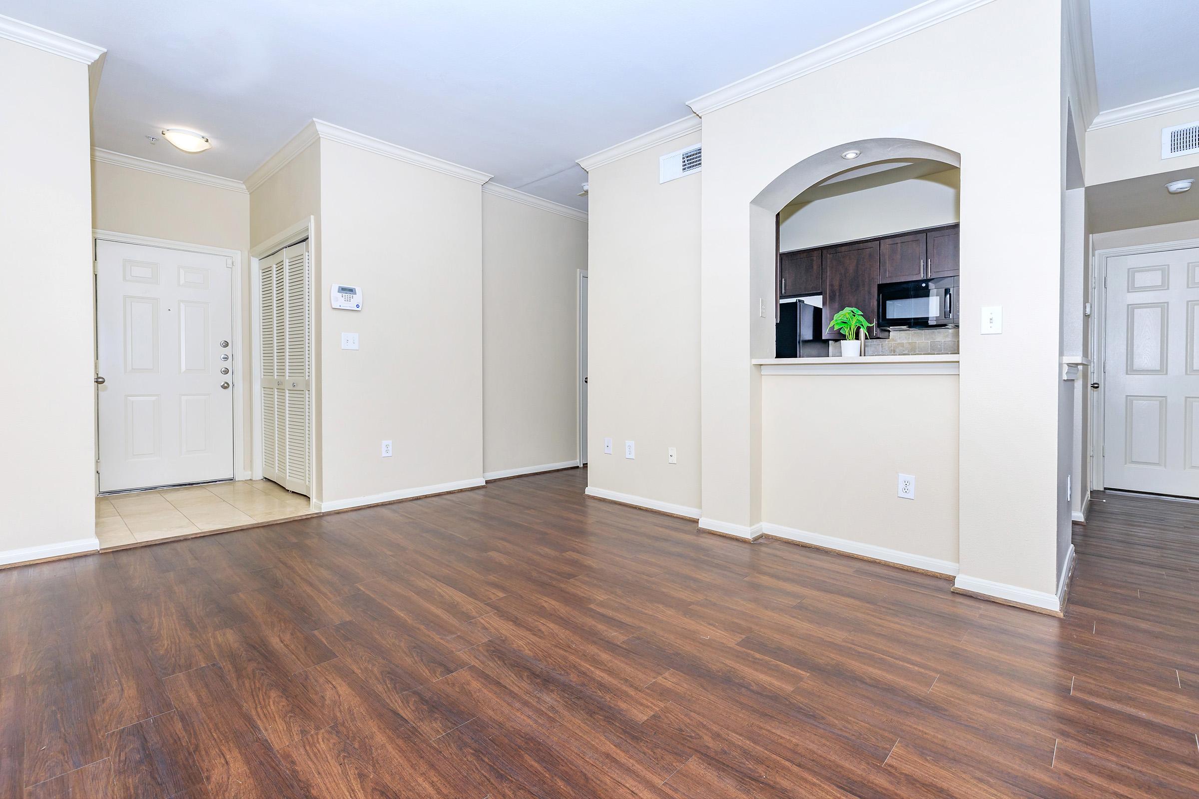 An empty living room showing a hardwood floor, beige walls, and a doorway leading to a kitchen area with dark cabinets. Two doors indicate entrances, and there is a small bar counter with a green plant on it. Natural light brightens the space, creating a welcoming atmosphere.