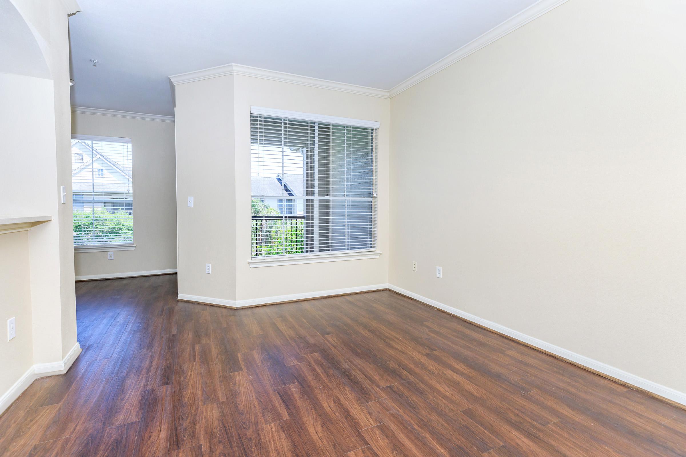 Empty interior of a room featuring light-colored walls, a window with blinds allowing natural light, and hardwood flooring. A corner of the room leads to another area, creating an open space layout.