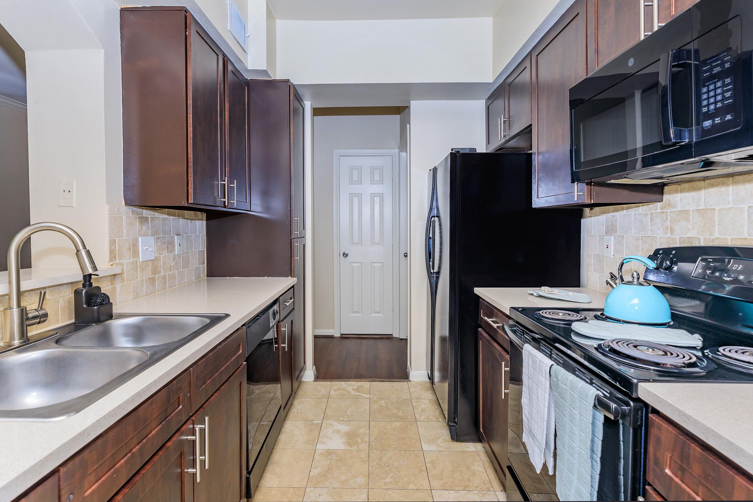 A modern kitchen featuring dark wood cabinets, stainless steel appliances, and light-colored countertops. The layout includes a double sink, an electric stove with an oven, a microwave overhead, and a refrigerator. The floor is tiled, and there is a door leading to another room in the background.