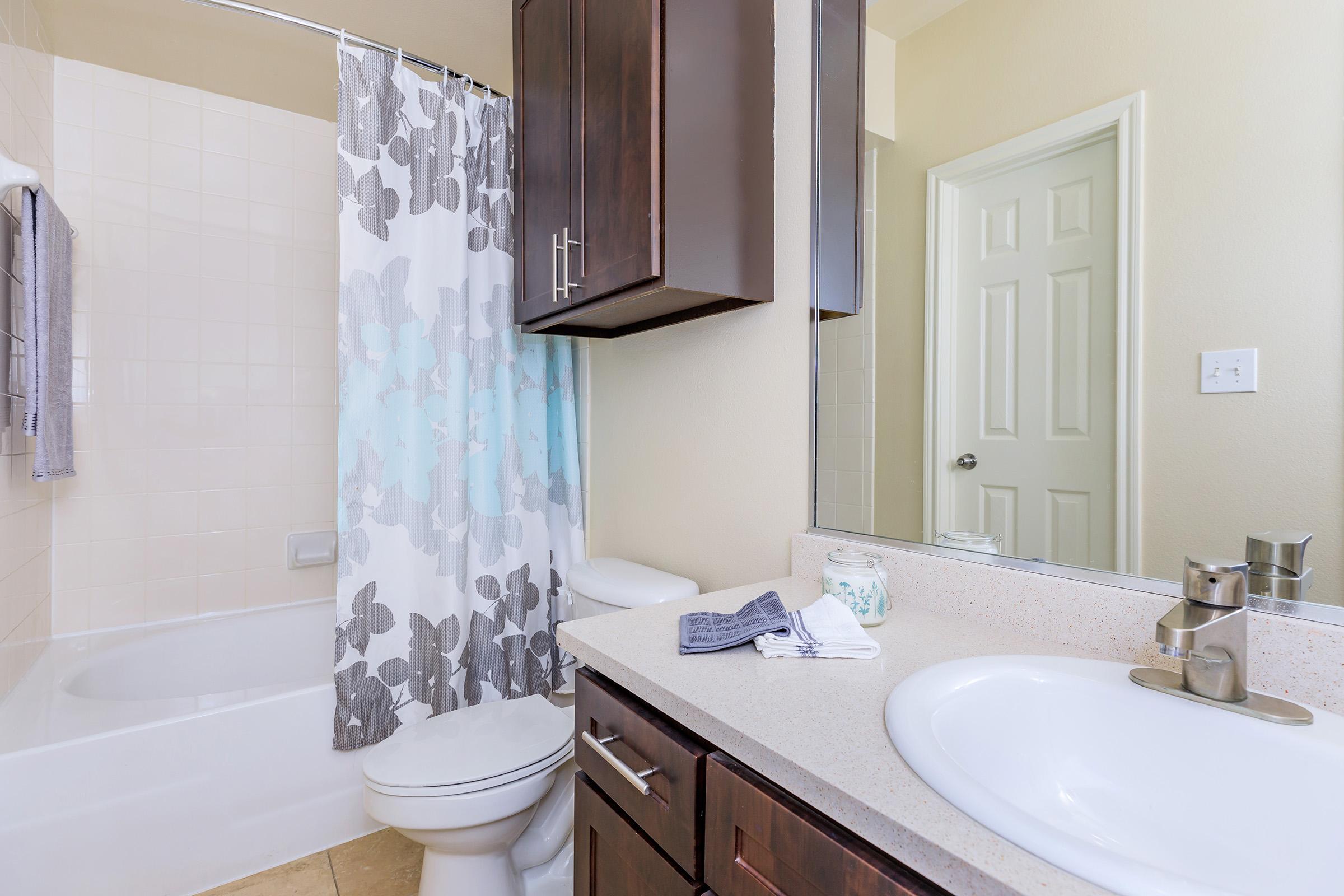 A modern bathroom featuring a white bathtub with a floral shower curtain, a wooden cabinet with a sink, a mirror, and a toilet. The walls are painted a light color, and there's a towel hanging on the wall. The space is clean and well-organized.