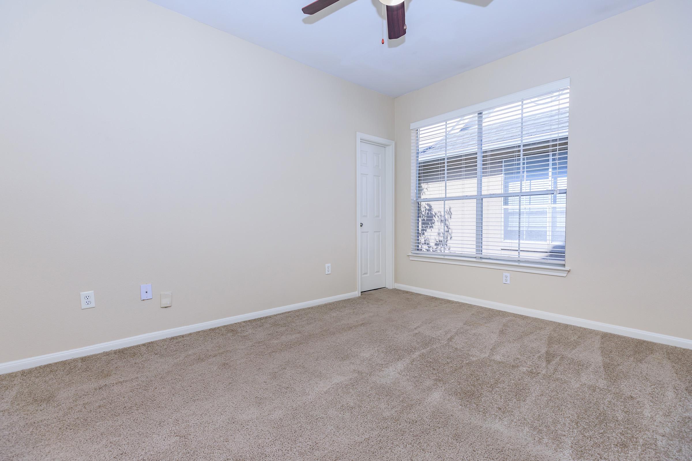 A clean, empty room with light beige walls and carpet. There is a window with blinds letting in natural light, and a white door on the left. The room has a ceiling fan and a simple, minimalist design, creating a spacious feel.