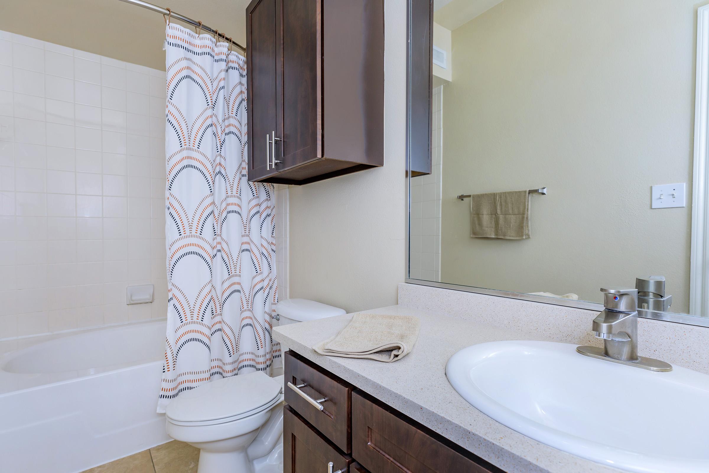 A clean and modern bathroom featuring a white bathtub with a shower, a patterned shower curtain, a dark wooden vanity with a sink, and a mirror. There’s a towel hanging on the wall and a hand towel placed on the countertop, with light-colored walls and tiled flooring.