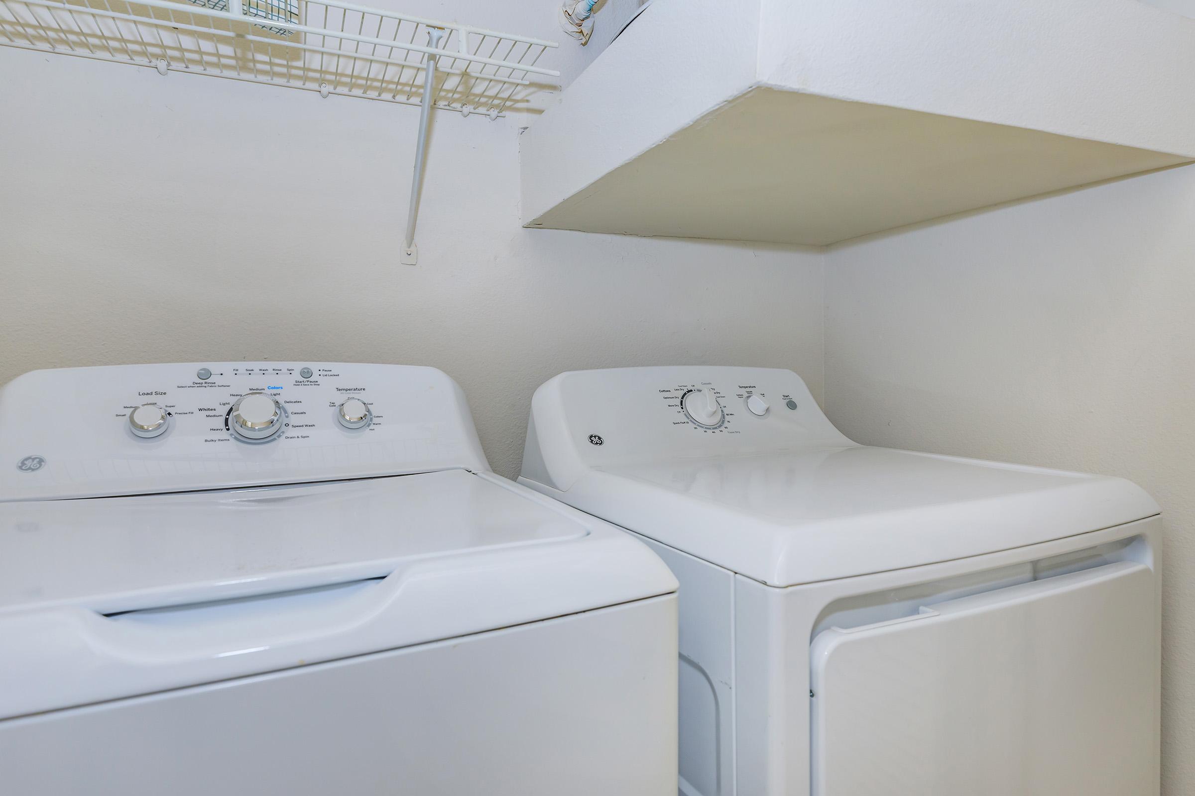 A white washing machine and dryer side by side in a laundry room. The machines have control dials on the front, and the room features a simple white wall and a shelf above the appliances.
