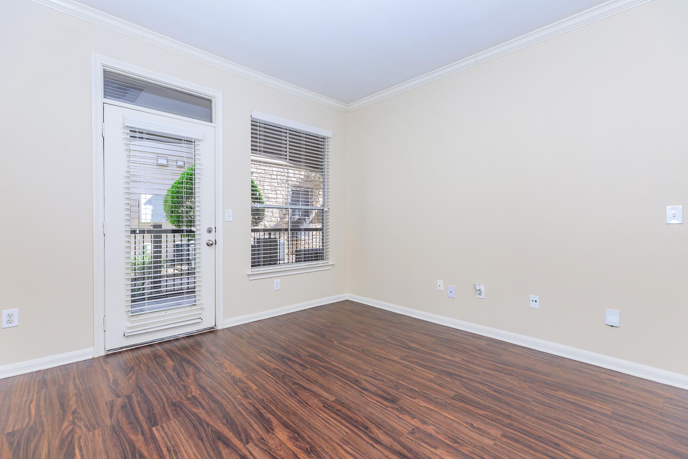 A spacious, empty room with light-colored walls and a hardwood floor. There's a door with glass panes leading outside and several windows with blinds letting in natural light. The area is clean and ready for furnishing, with minimal decor and electrical outlets visible.