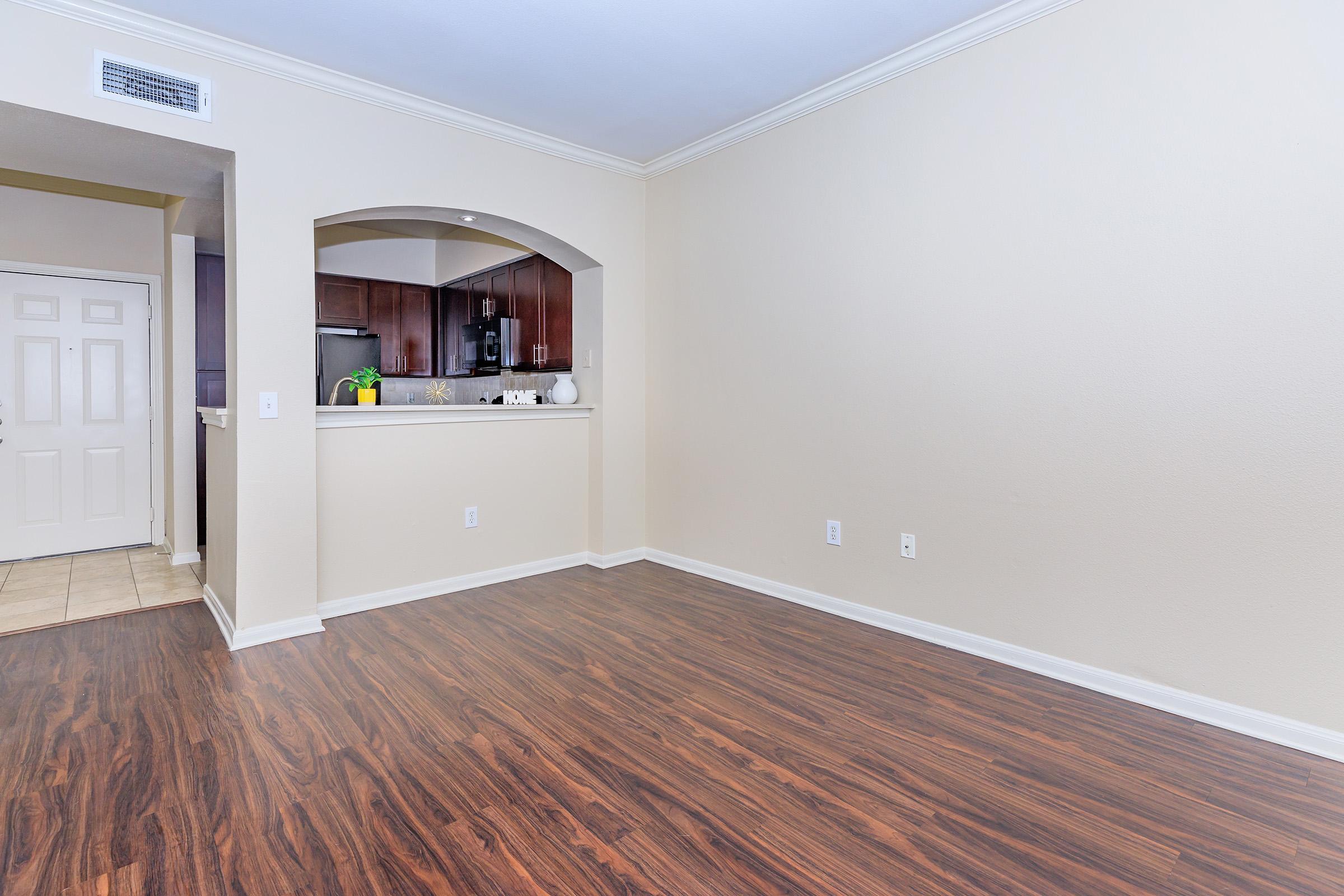A spacious, empty room with light wood laminate flooring and neutral-colored walls. A small kitchen with dark wood cabinets is visible through an archway. A door leads to another room. The area is well-lit with natural light, creating a welcoming atmosphere.