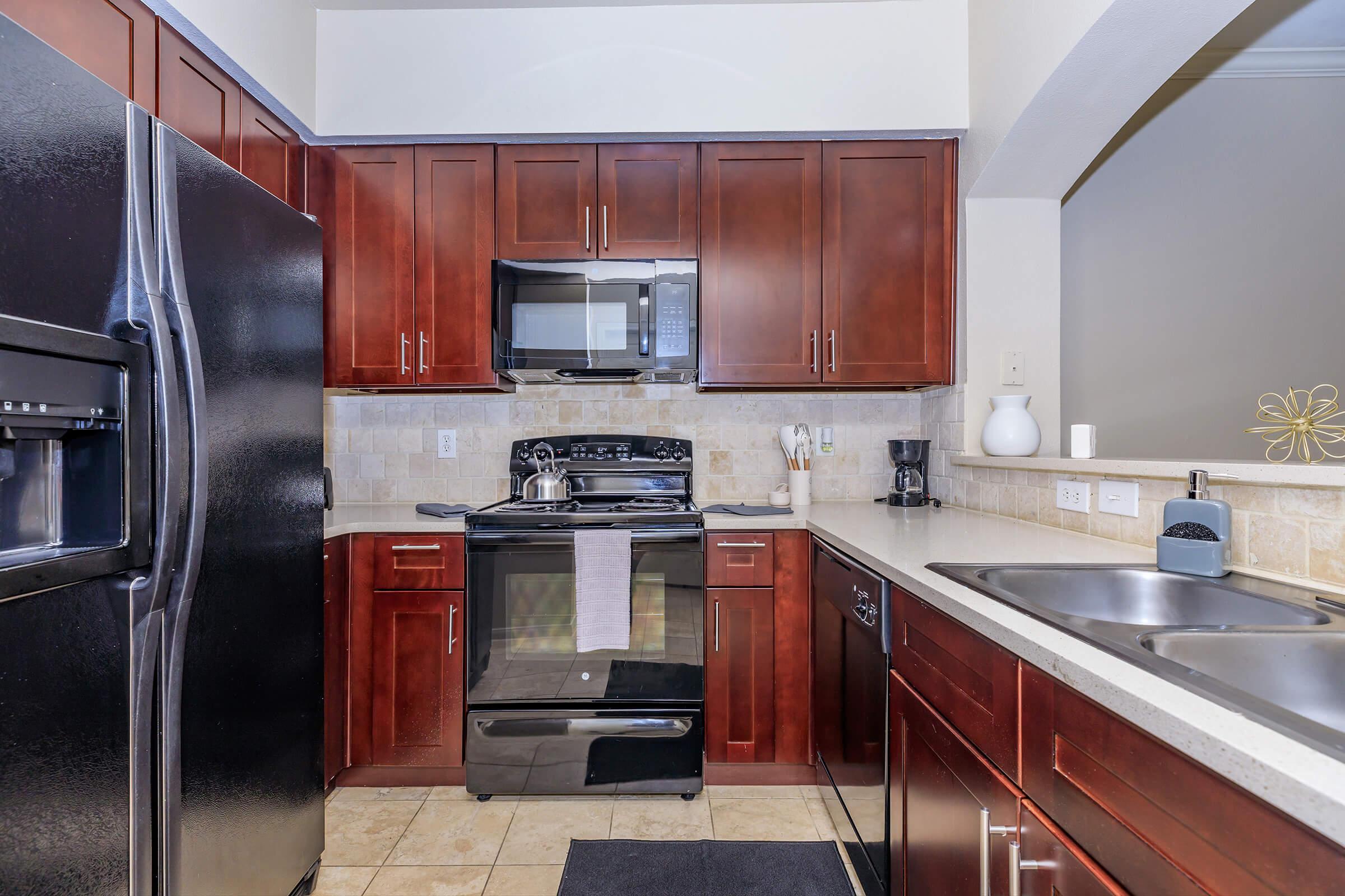 Modern kitchen featuring dark wood cabinetry, stainless steel appliances, including a refrigerator, microwave, and stove. The countertop is light-colored, with a double sink and a dark mat on the floor. The walls are adorned with light tiles, creating a warm and inviting atmosphere.