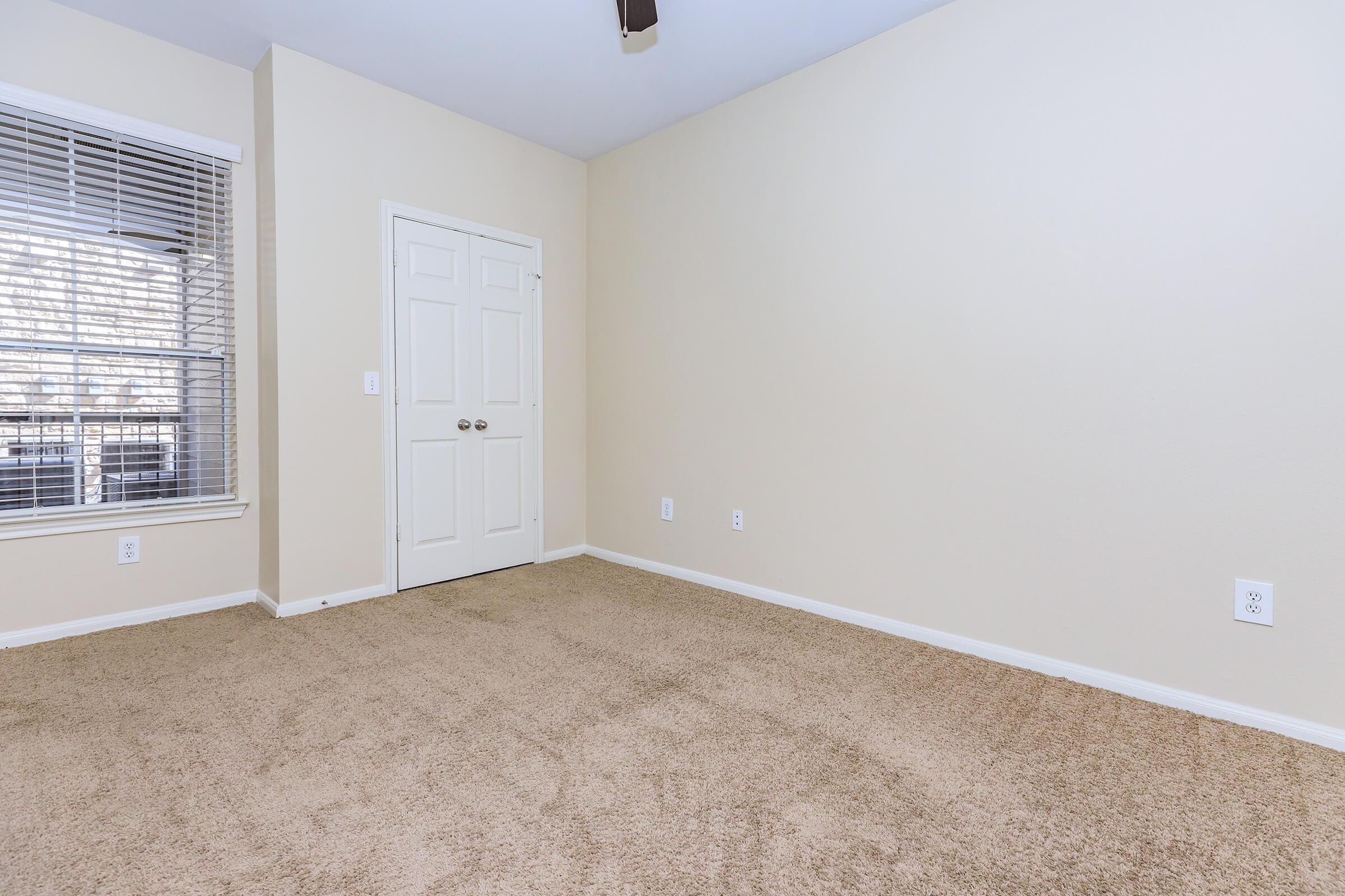 A spacious, empty room with beige walls and carpet, featuring a single closed door on the right and a window with blinds on the left. The room is well-lit and has a ceiling fan, giving it a warm and inviting atmosphere.