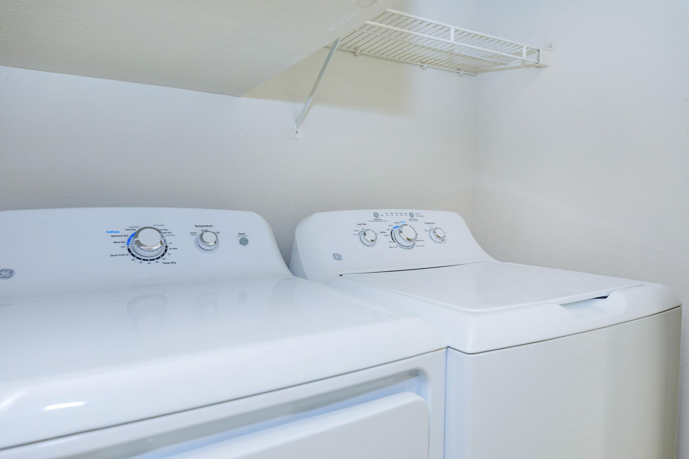 Two white appliances in a laundry area: a top-loading washing machine on the left and a dryer on the right. Above the machines, there is a wall-mounted shelf for storage. The wall is painted a light color, creating a clean and organized look.