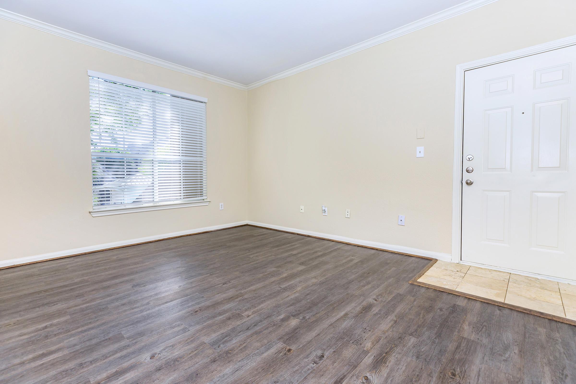 A spacious, empty room with light-colored walls and hardwood flooring. A large window with white blinds allows natural light to enter. The room features a white door on the right side and tile flooring at the entrance, creating a welcoming atmosphere.