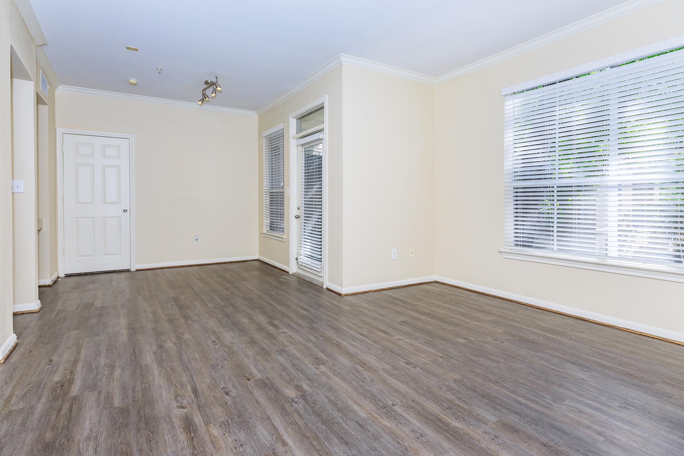 A spacious, empty room with light-colored walls and hardwood flooring. There are large windows letting in natural light, and a door on one wall. The room is well-lit and clean, creating a bright and open atmosphere.