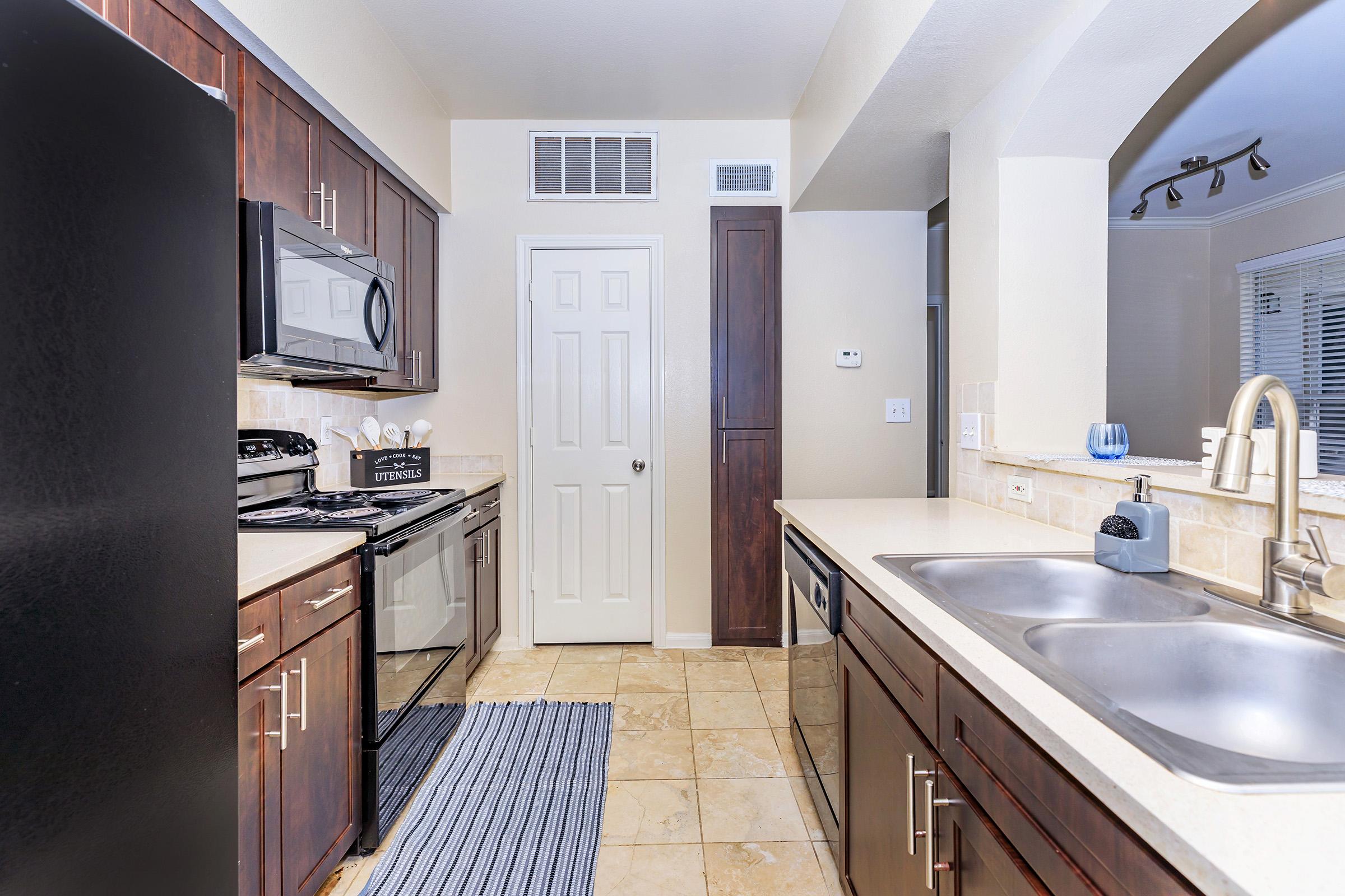 Modern kitchen featuring dark wood cabinets, stainless steel appliances, and beige countertops. The layout includes a refrigerator, microwave, oven, and dual sink. A blue and white striped rug is laid on the floor, and a door leads to another room, with a wall-mounted thermostat visible.
