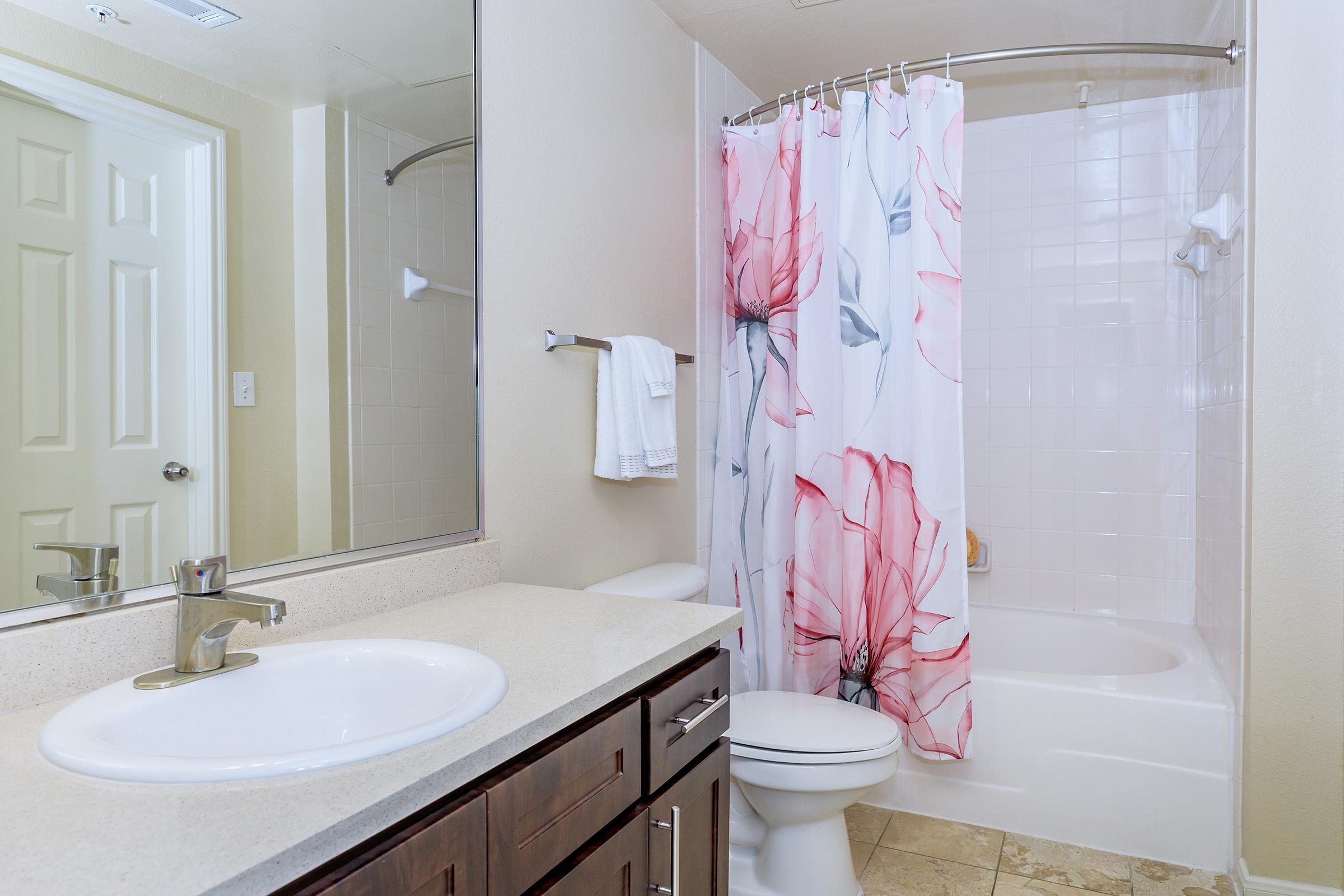 A bathroom featuring a white bathtub and shower with a floral-patterned shower curtain. A rectangular mirror above the sink reflects the space. The sink has a modern faucet and sits on a beige countertop, while a towel is hanging nearby. The walls are light-colored, creating a bright atmosphere.