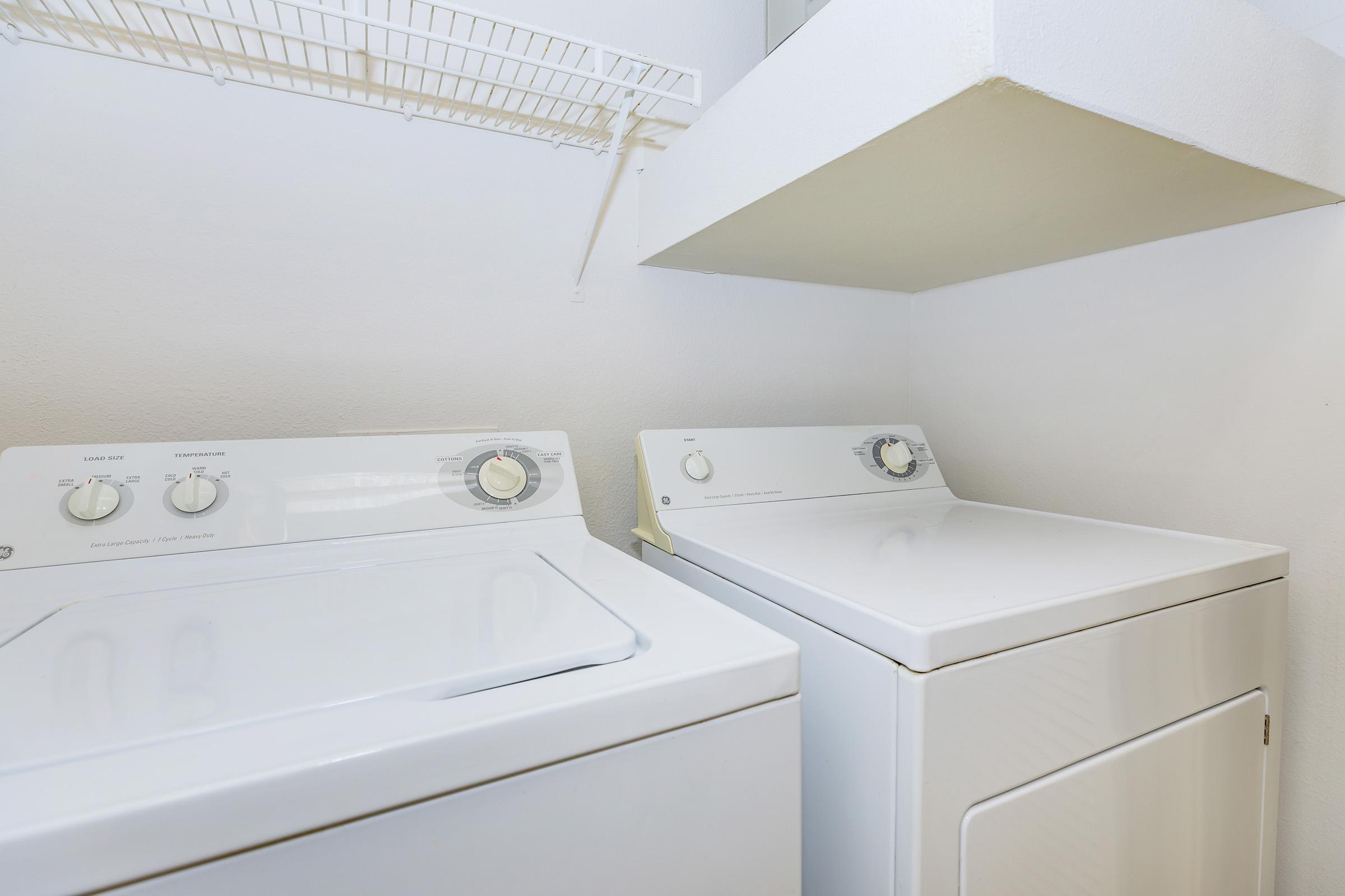 A clean laundry area featuring a washing machine and a dryer beside each other. The wall is painted white, and there is a shelf above the appliances for storage.