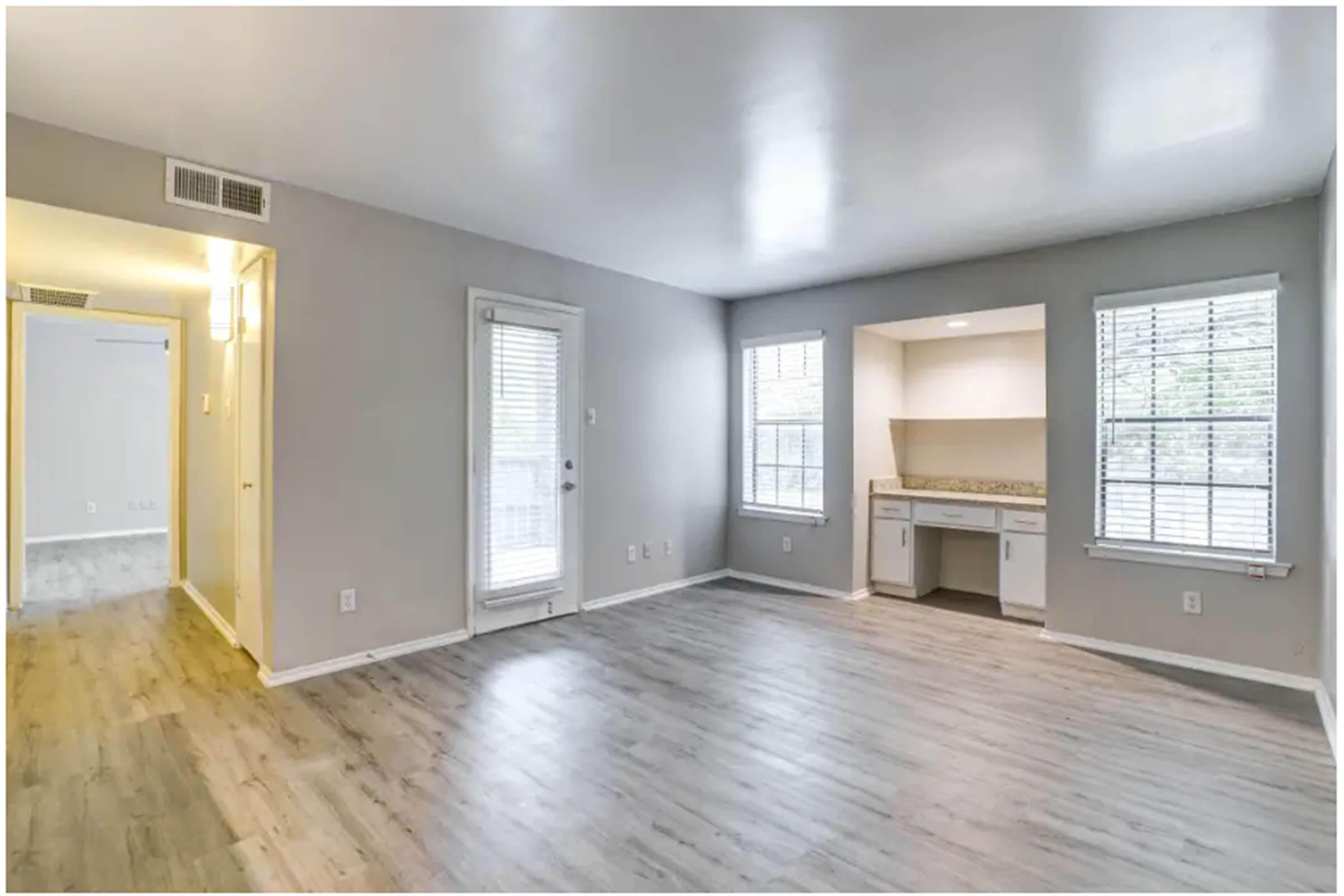 A spacious, empty room with light gray walls and wooden flooring. There are two windows with blinds letting in natural light, and a small built-in desk area with countertop. A door leads to an outside area, and another doorway opens to another room in the background.