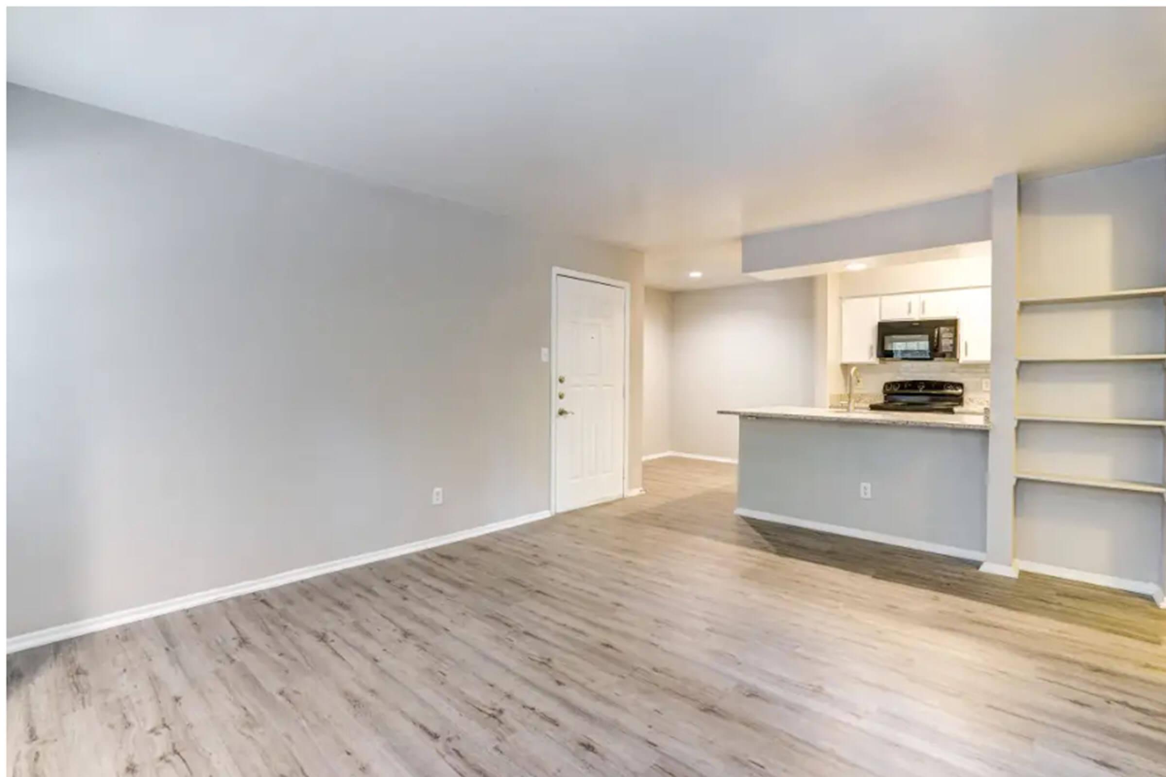 Spacious, unfurnished living room with light gray walls and wooden flooring. A doorway leads to an entrance, and there is a kitchen area featuring modern appliances and a breakfast bar with seating. Natural light brightens the space from a nearby source. Shelf unit against the wall adds to the decor.