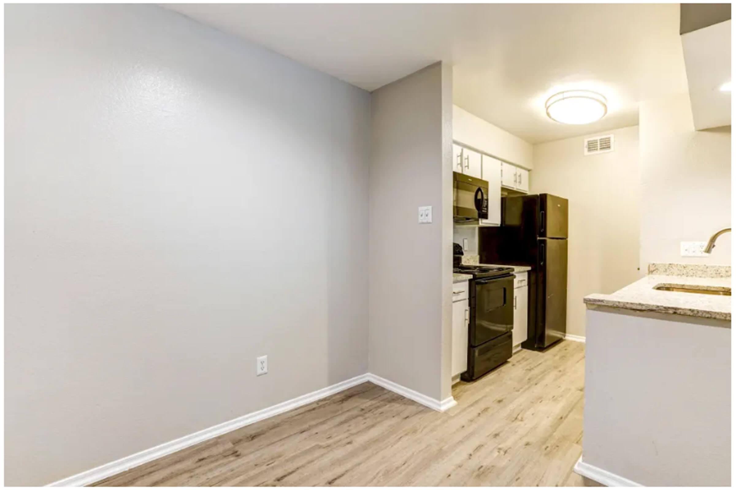 A modern kitchen featuring light gray walls, wooden flooring, and stainless steel appliances. The space includes a black stove, microwave, and refrigerator. A granite countertop separates the kitchen from a small dining area, illuminated by a round ceiling light.