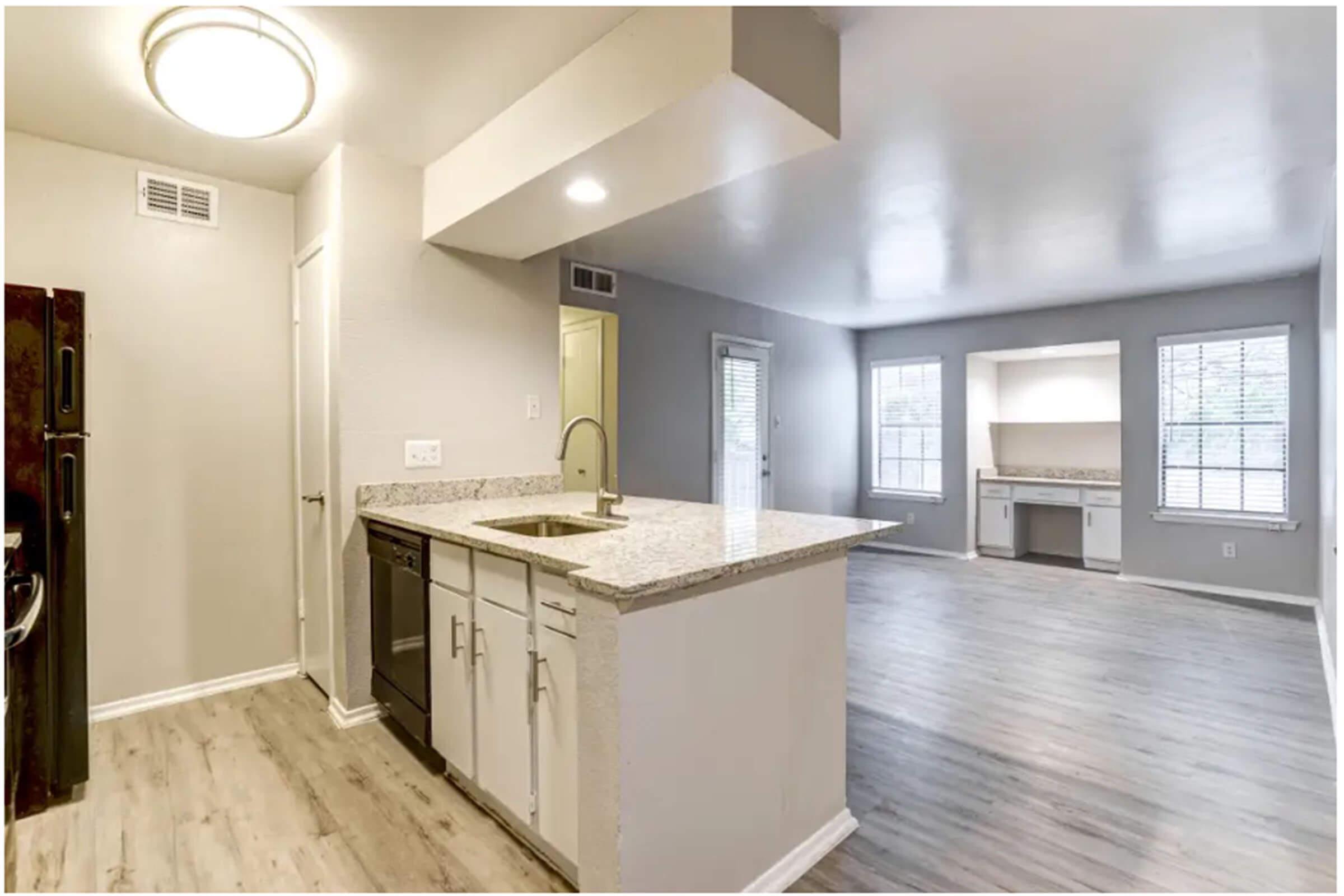 A modern kitchen with granite countertops and stainless steel appliances, including a sink, dishwasher, and refrigerator. The open layout leads into a well-lit living area with large windows and light-colored walls. Wood-like flooring runs throughout the space, creating a bright and inviting atmosphere.