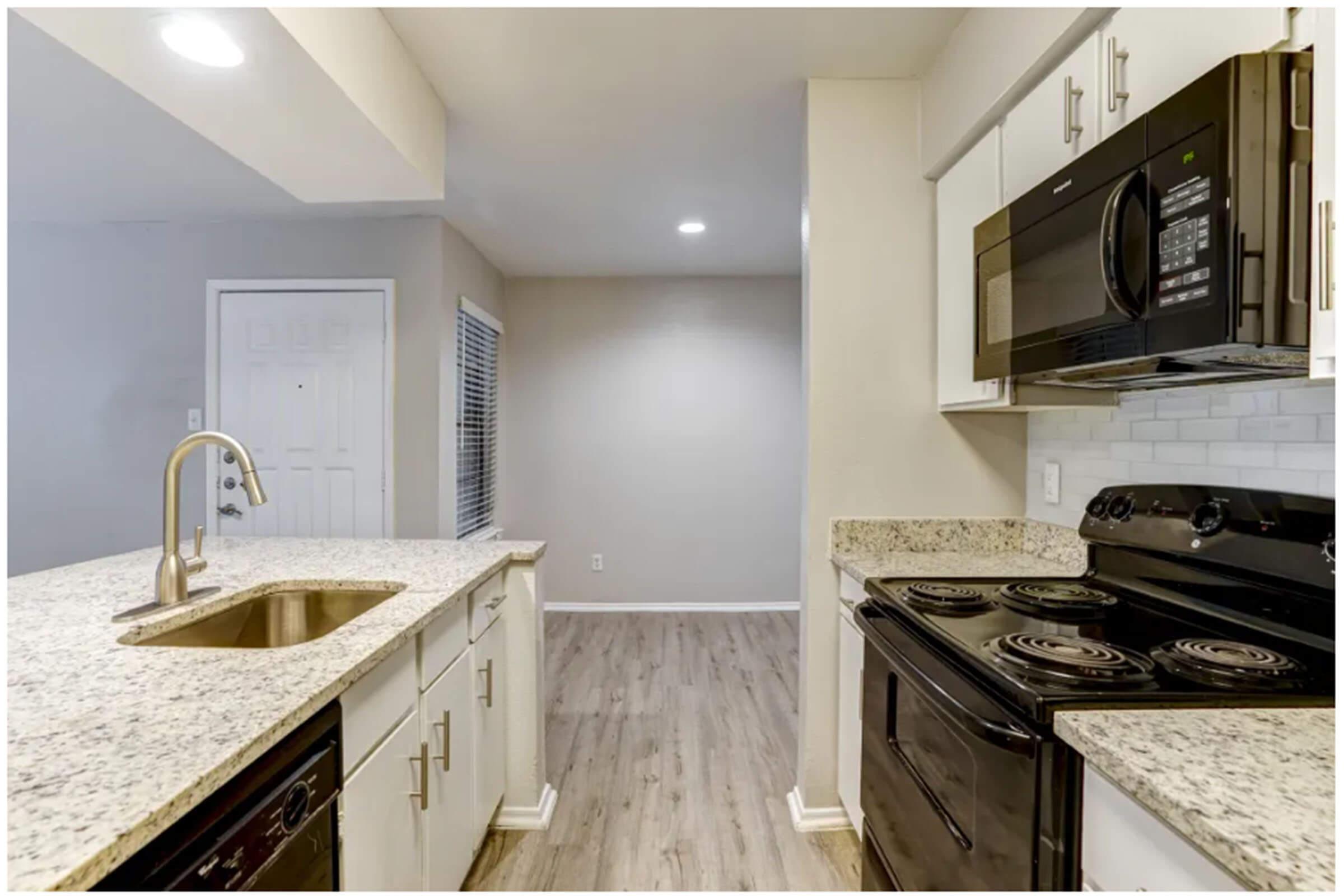 Modern kitchen featuring white cabinets, a granite countertop with a sink, a black microwave, and a black stove. The kitchen is well-lit with recessed lighting, and there is an adjacent living area visible. The flooring appears to be laminate wood.