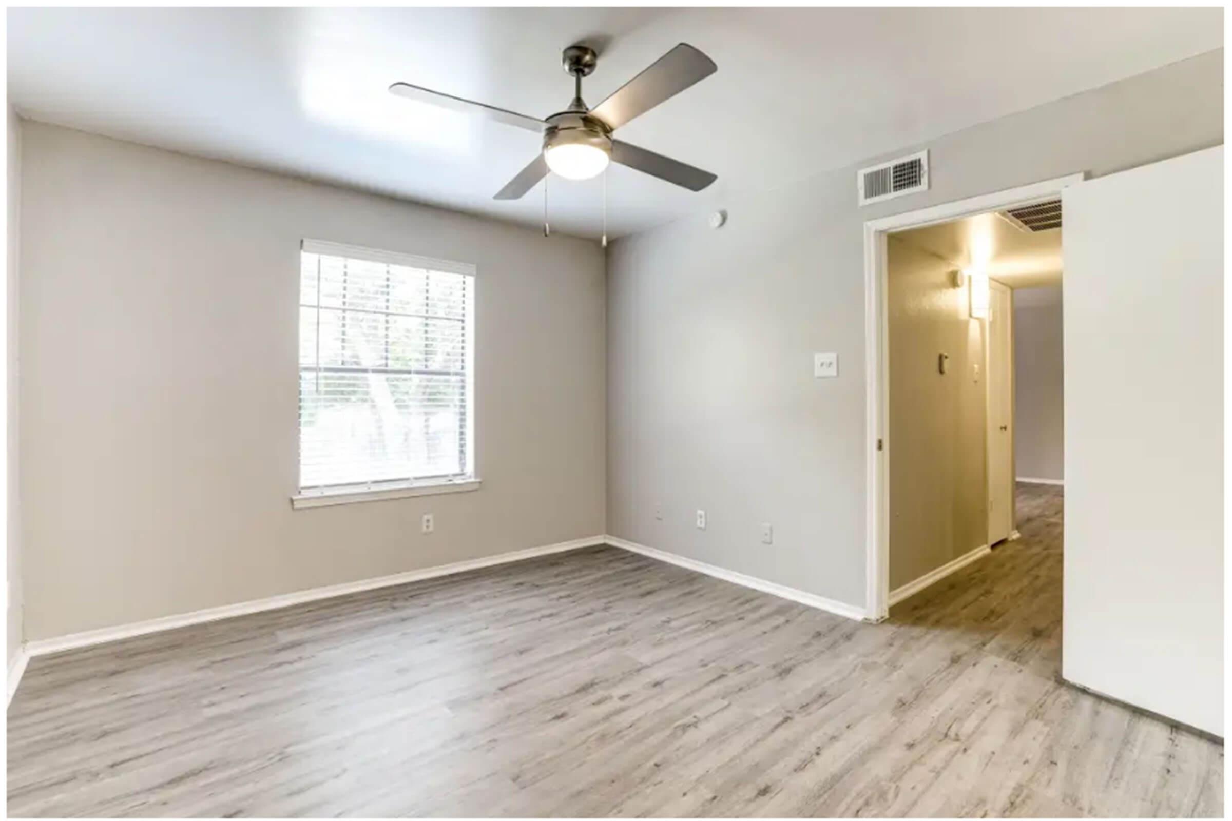 A well-lit, empty room featuring light gray walls, laminate wood flooring, and a ceiling fan. There's a window with white blinds on the left and an open door on the right leading to a hallway. The space appears clean and ready for furnishing.