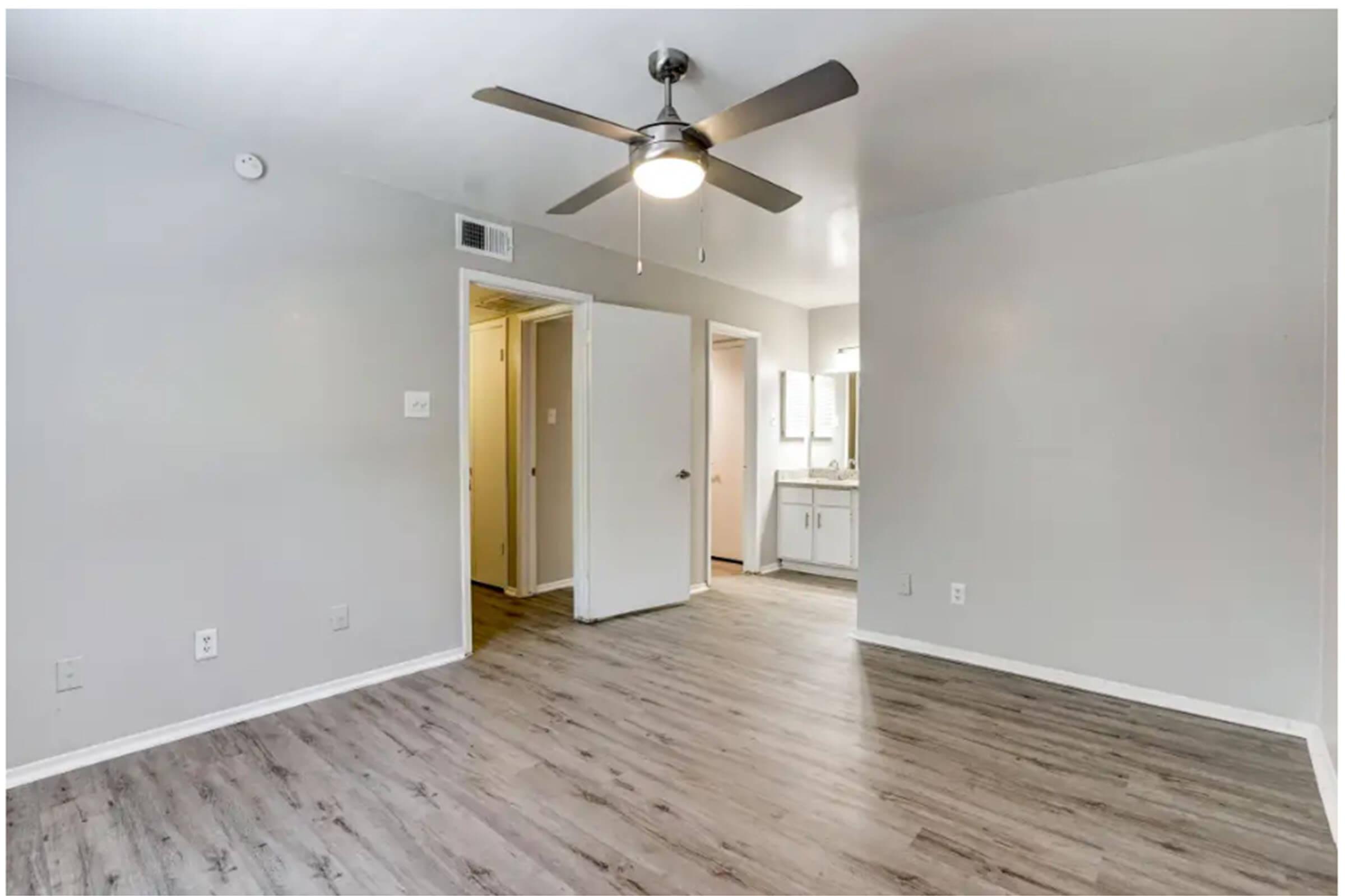 View of a light-filled room with a ceiling fan, neutral walls, and hardwood-style flooring. A partially open door leads to a hallway, while another door opens to a bathroom area with a vanity. The minimalist design creates a modern, spacious feel.