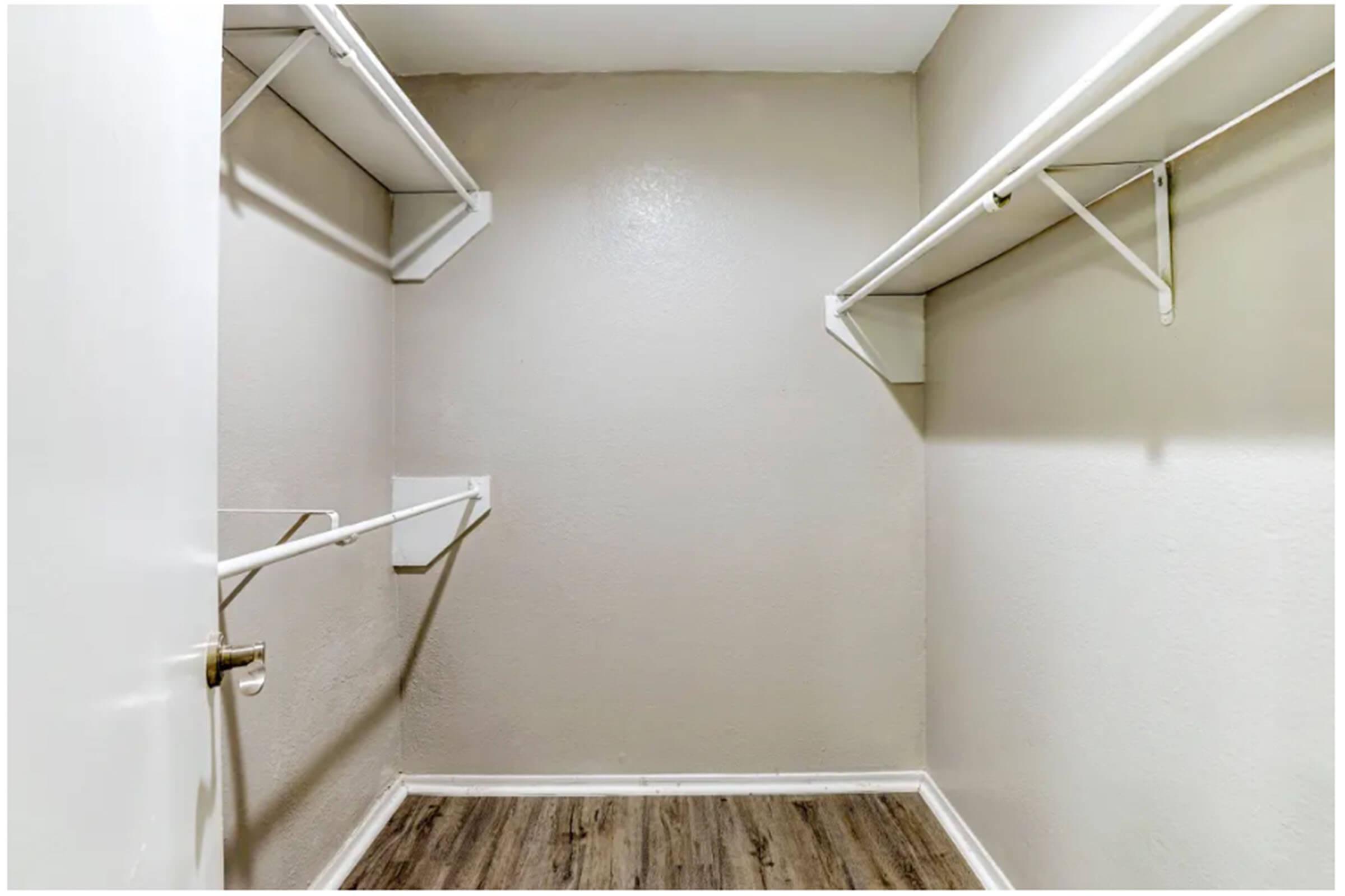 Empty closet with light-colored walls and a wooden floor. Two wall-mounted shelves are positioned high on each side, and there is a single clothing rod attached to one side. The space is well-lit, creating an open and airy feel despite the lack of furnishings.