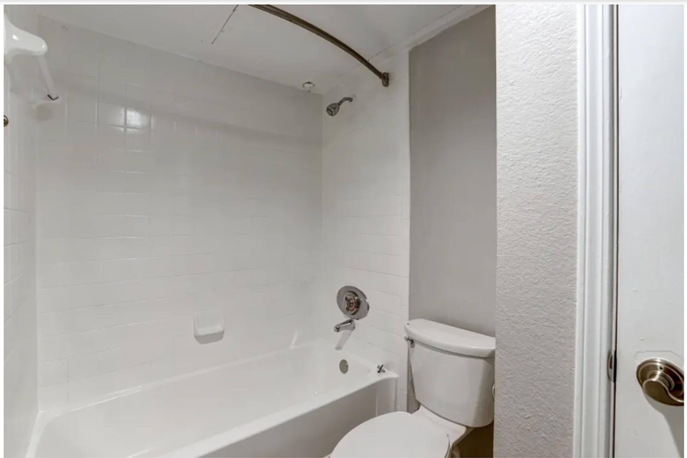 A clean, modern bathroom featuring a white bathtub with a showerhead above, white tiled walls, and a light gray wall. There is a toilet beside the bathtub and a door knob visible in the foreground, suggesting an entrance to the room. The overall atmosphere is bright and minimalistic.