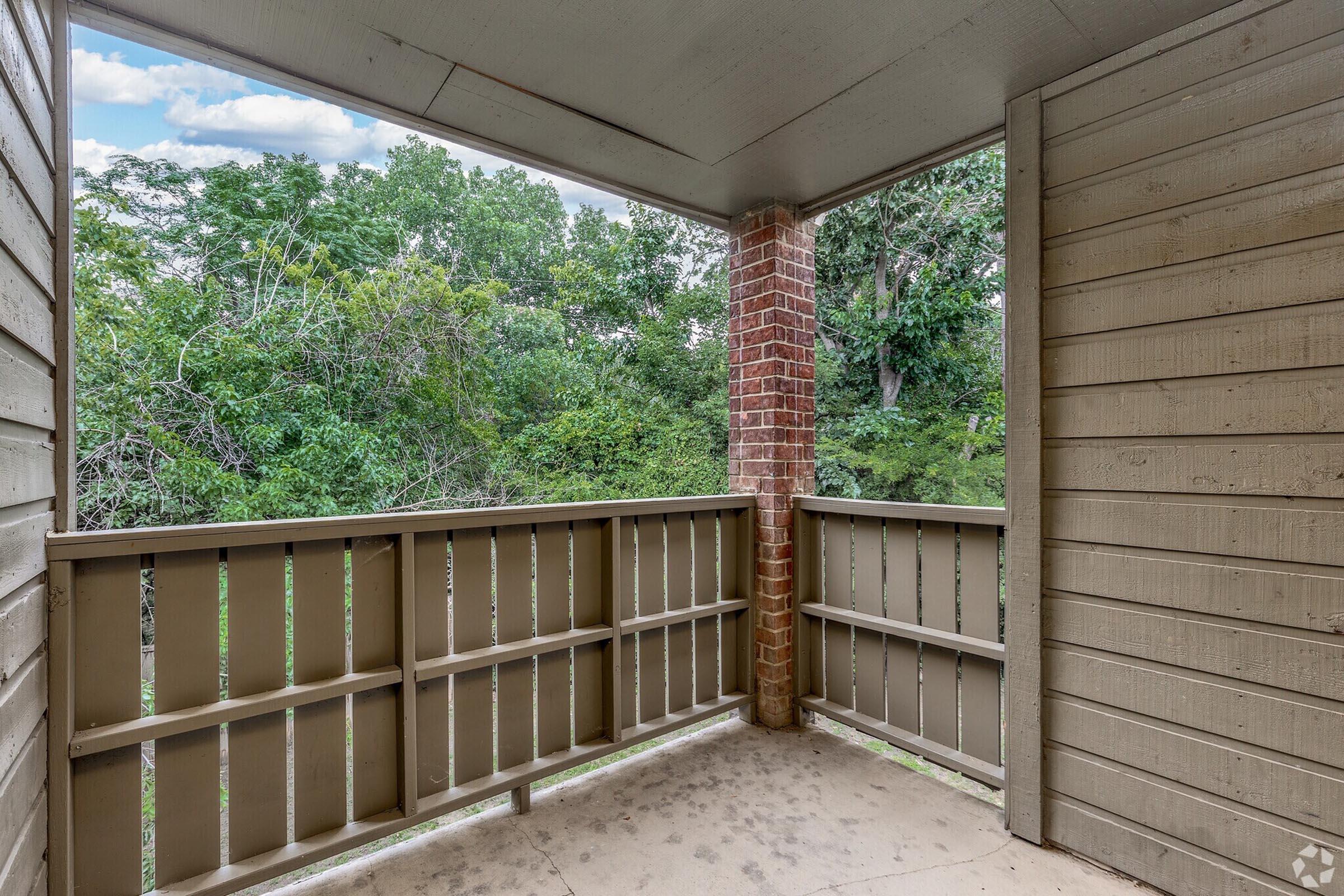 View from a balcony overlooking a green landscape. The balcony features rustic wooden railings and a ceiling, surrounded by trees and foliage in the background. The scene conveys a peaceful outdoor setting, ideal for relaxation.