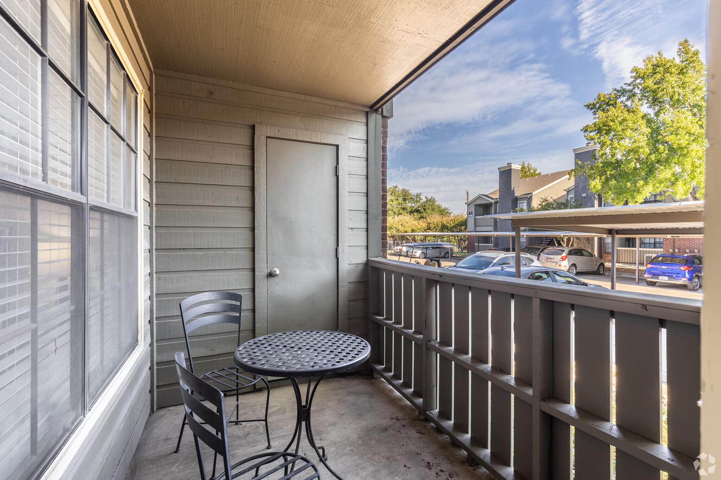 A small balcony featuring a round black metal table with two chairs, a door leading inside, and a view of parked cars and greenery outside. The walls are painted in a light tone with large windows allowing natural light. The sky is partly cloudy, creating a serene atmosphere.