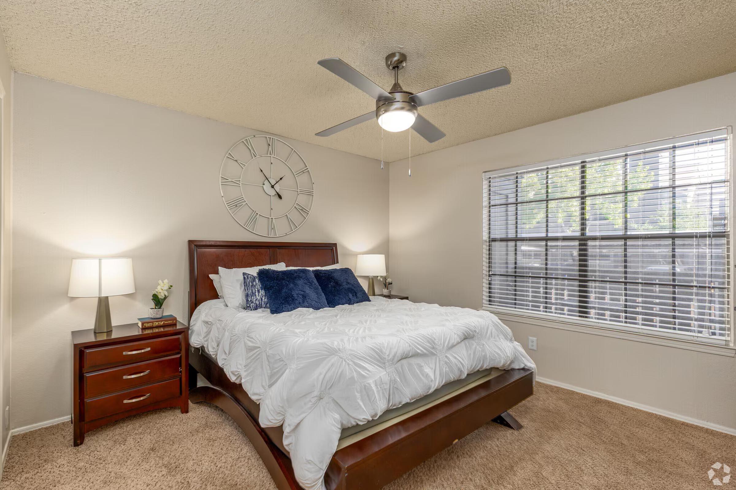 A cozy bedroom featuring a large bed with white bedding and blue accent pillows. There are two bedside tables with lamps, a clock on the wall, and a window with blinds allowing natural light to enter. A ceiling fan is seen above, adding to the comfortable ambiance.