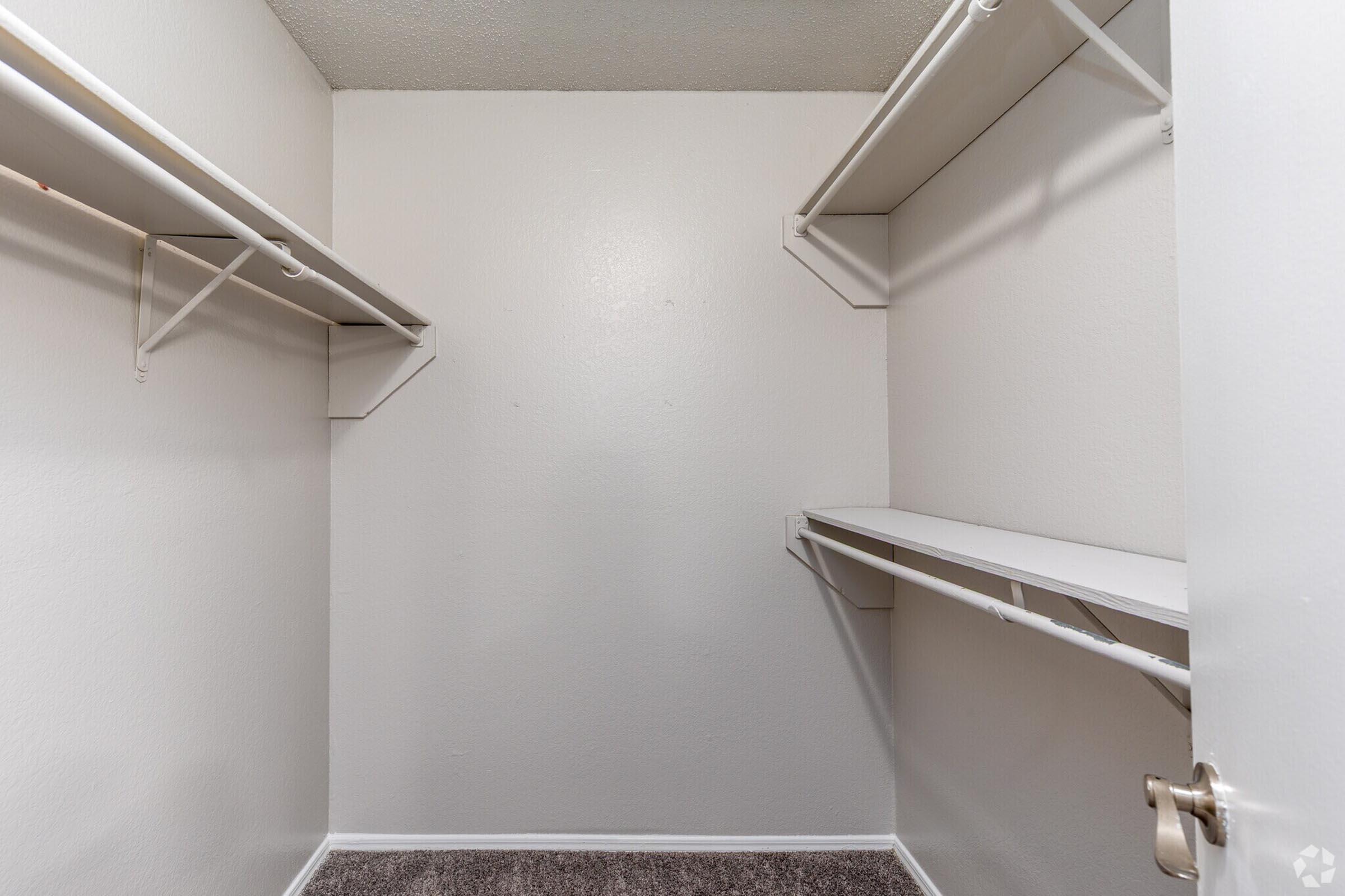 A clean, empty closet featuring white walls and a carpeted floor. It has two shelves mounted on the walls and a long shelf running along one side. The closet is well-lit and spacious, providing ample storage potential. The door is partially open, revealing the uncluttered interior.