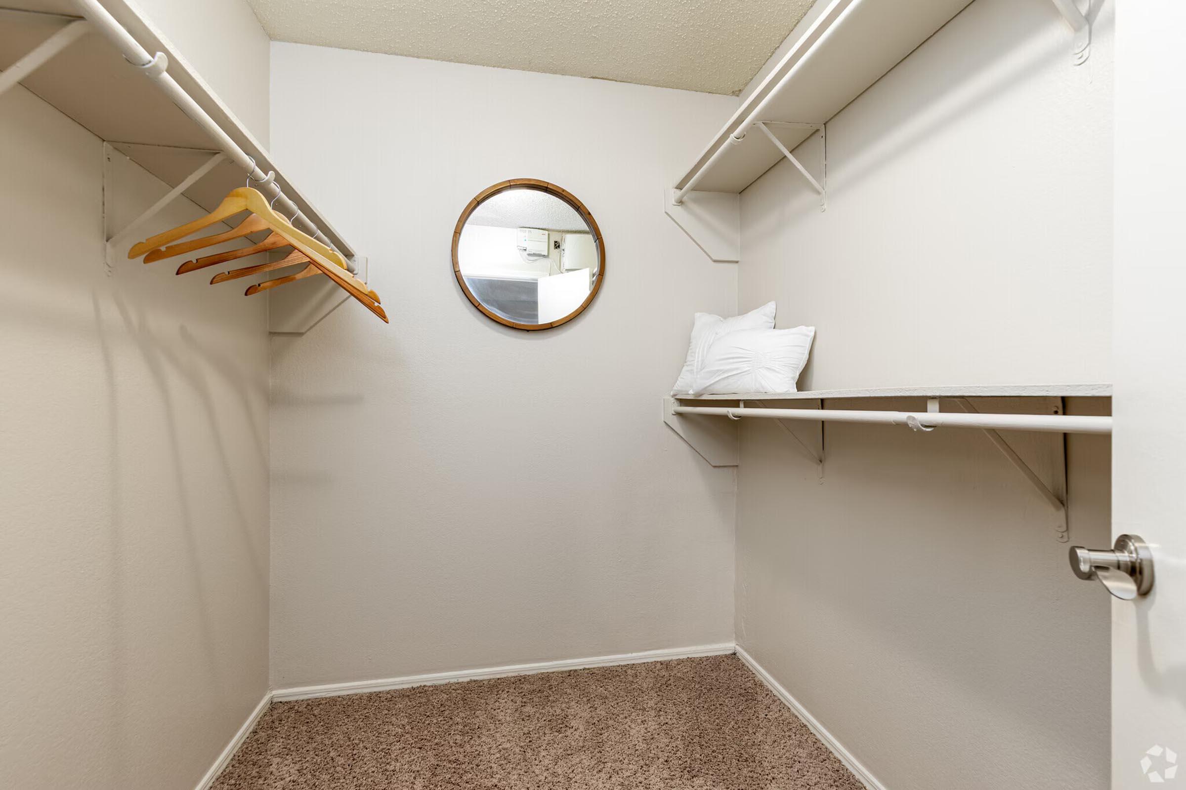 A small, empty closet featuring two shelves, a round mirror on the wall, and a couple of wooden hangers. The floor is covered in light brown carpet, and the walls are painted white. The space is well-lit, providing a clean and minimalist look.