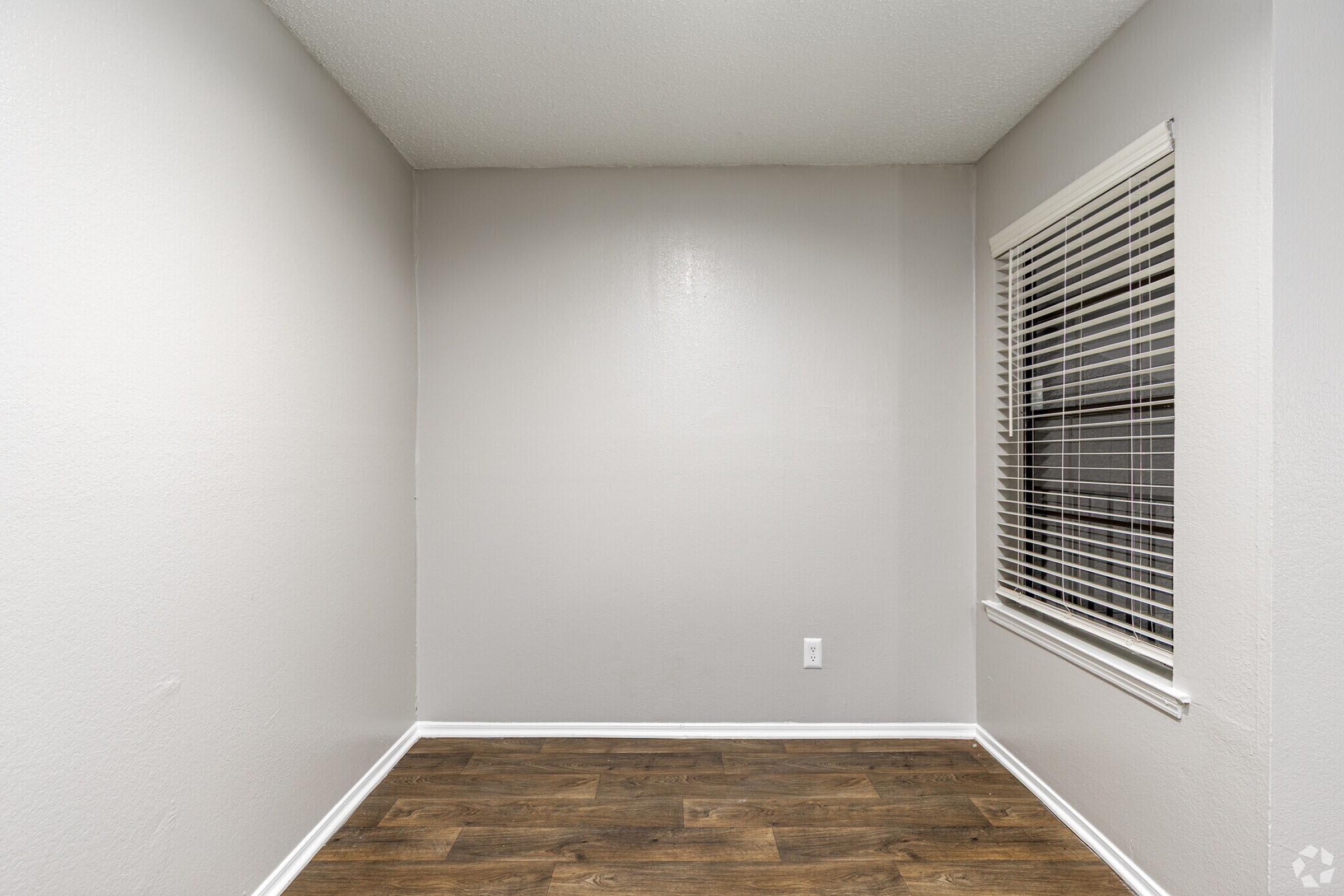 Empty room with light gray walls and a hardwood floor. A single window with white blinds is located on one wall, creating a bright, minimalist space. The corner of the room is visible, emphasizing its spaciousness and simplicity. No furniture or decorations are present.