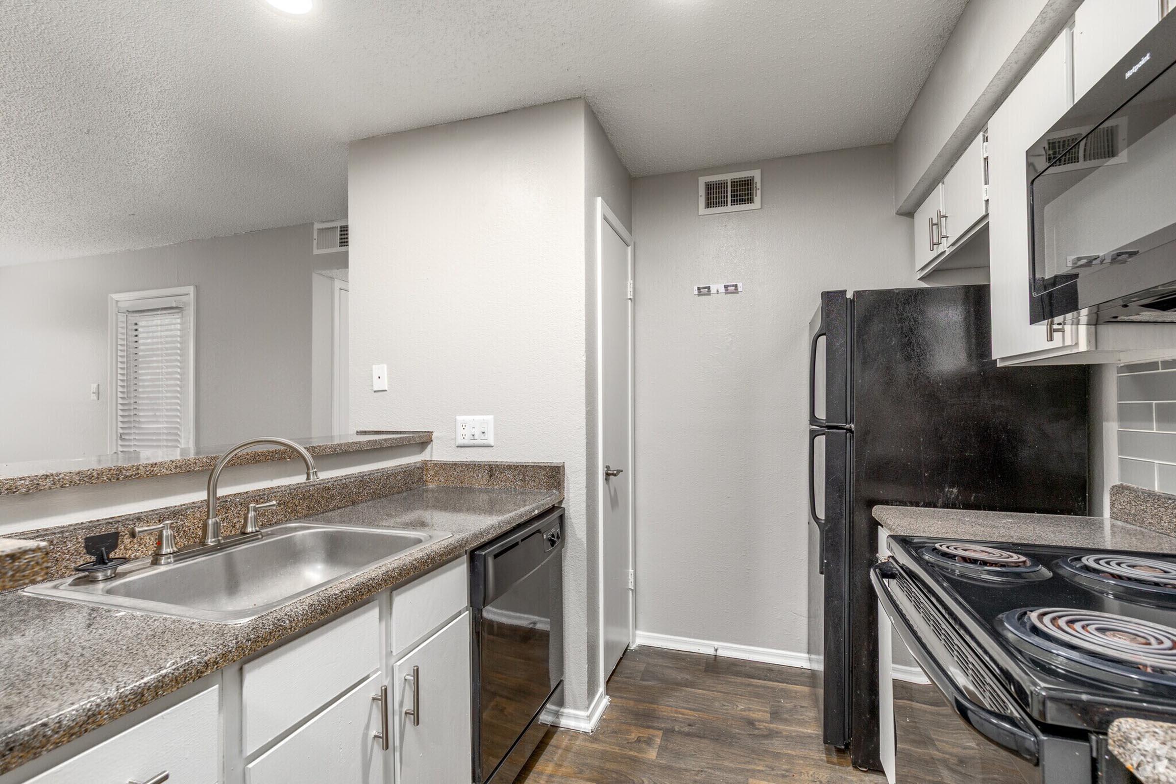 A modern kitchen featuring granite countertops, stainless steel sink, black appliances including a refrigerator and stove, and white cabinetry. The walls are painted a light gray, and there are wooden floors. The space is well-lit with overhead lighting, giving a clean and contemporary look.