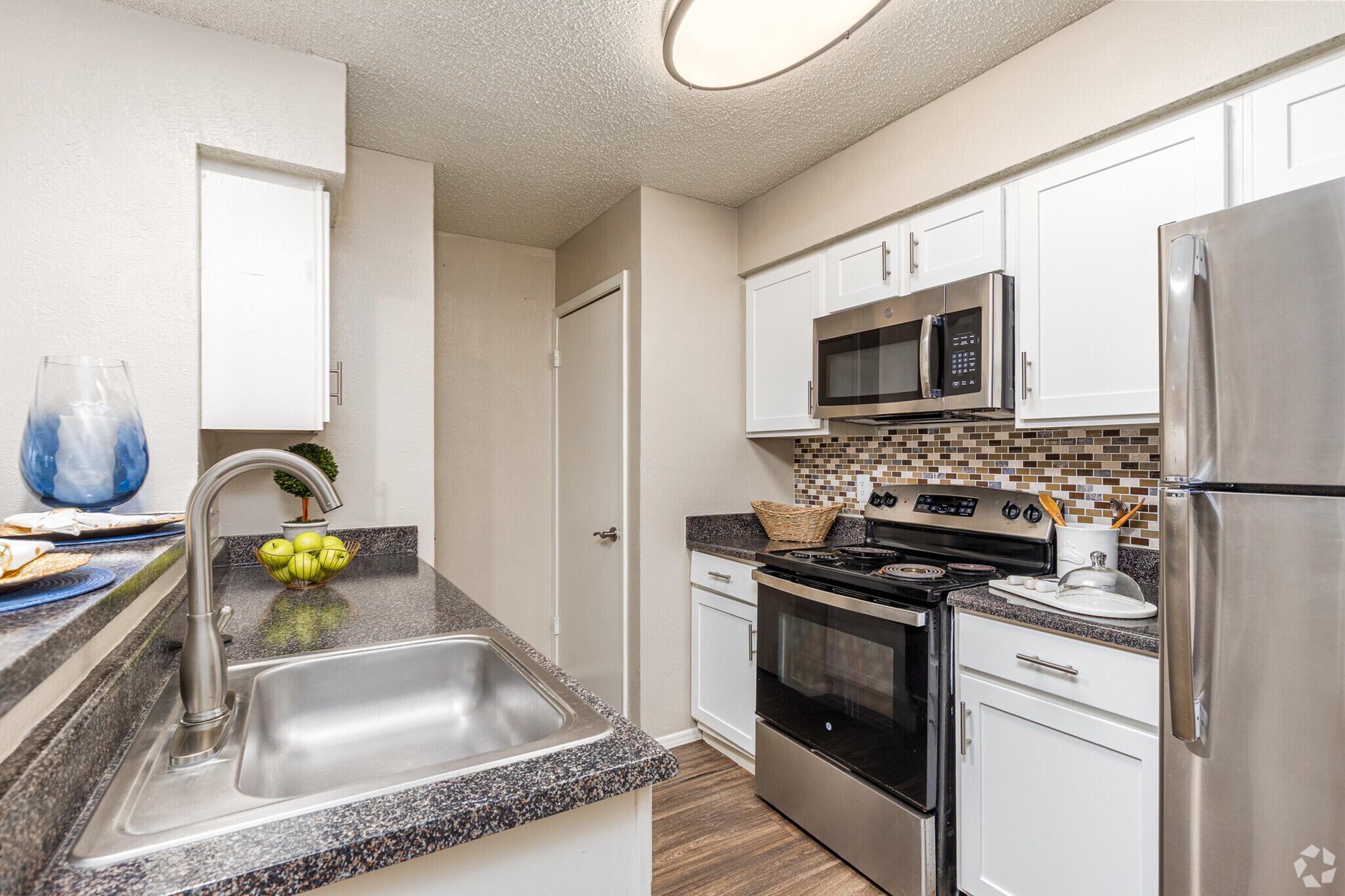 A modern kitchen featuring white cabinetry, a stainless steel refrigerator, a microwave, and a black stove. The counter is dark granite, and there's a stainless steel sink. Decorative items, including a blue vase and a bowl of fruit, add a touch of color. The walls are light-colored, creating a bright and inviting atmosphere.