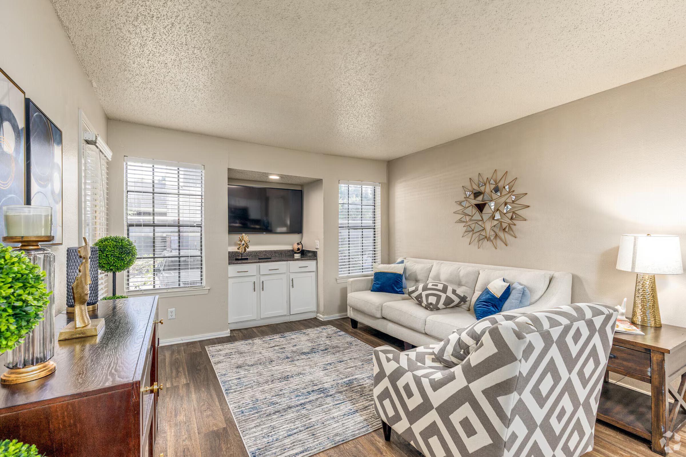 A cozy living room featuring a light-colored sofa with blue accent pillows, a patterned armchair, a small bar area with a flat-screen TV, and decorative items. Natural light filters through the window blinds, highlighting a stylish rug and a decorative mirror on the wall.