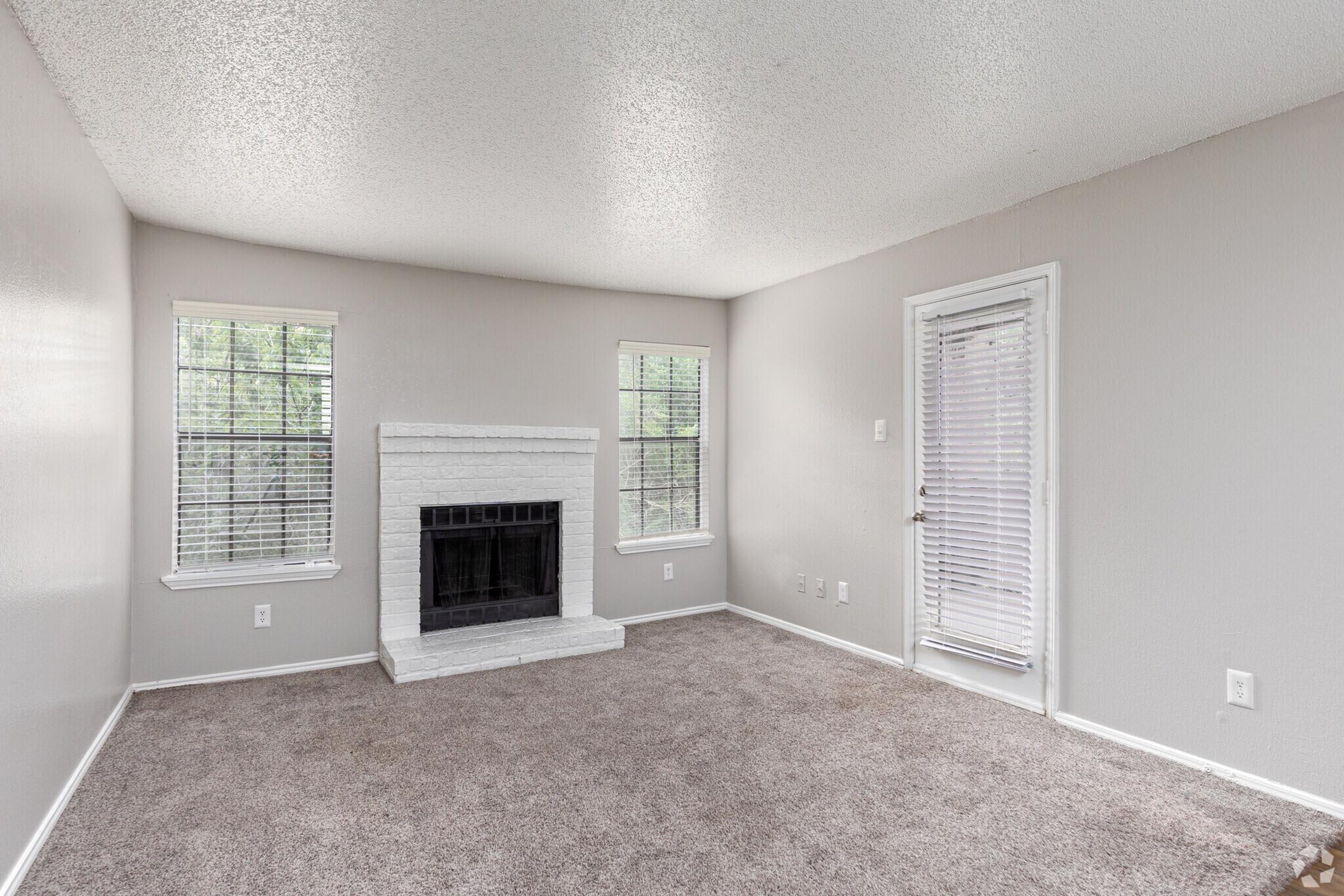A spacious living room featuring light gray walls and carpet, a white brick fireplace as a focal point, and large windows allowing natural light. A door leads outside, and there are no furnishings, creating a minimalist and open atmosphere.