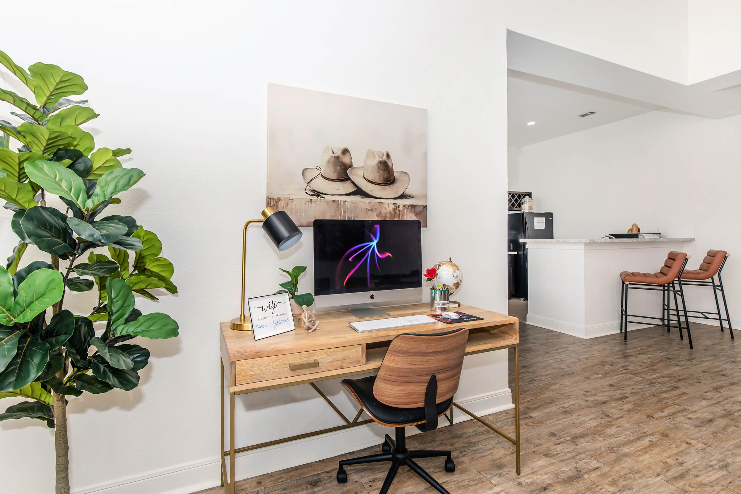 A modern workspace featuring a wooden desk with a computer and decorative items, a potted plant beside it, and a large framed artwork of two hats on the wall. In the background, a kitchen area with bar stools is visible. The overall ambiance is clean and inviting.