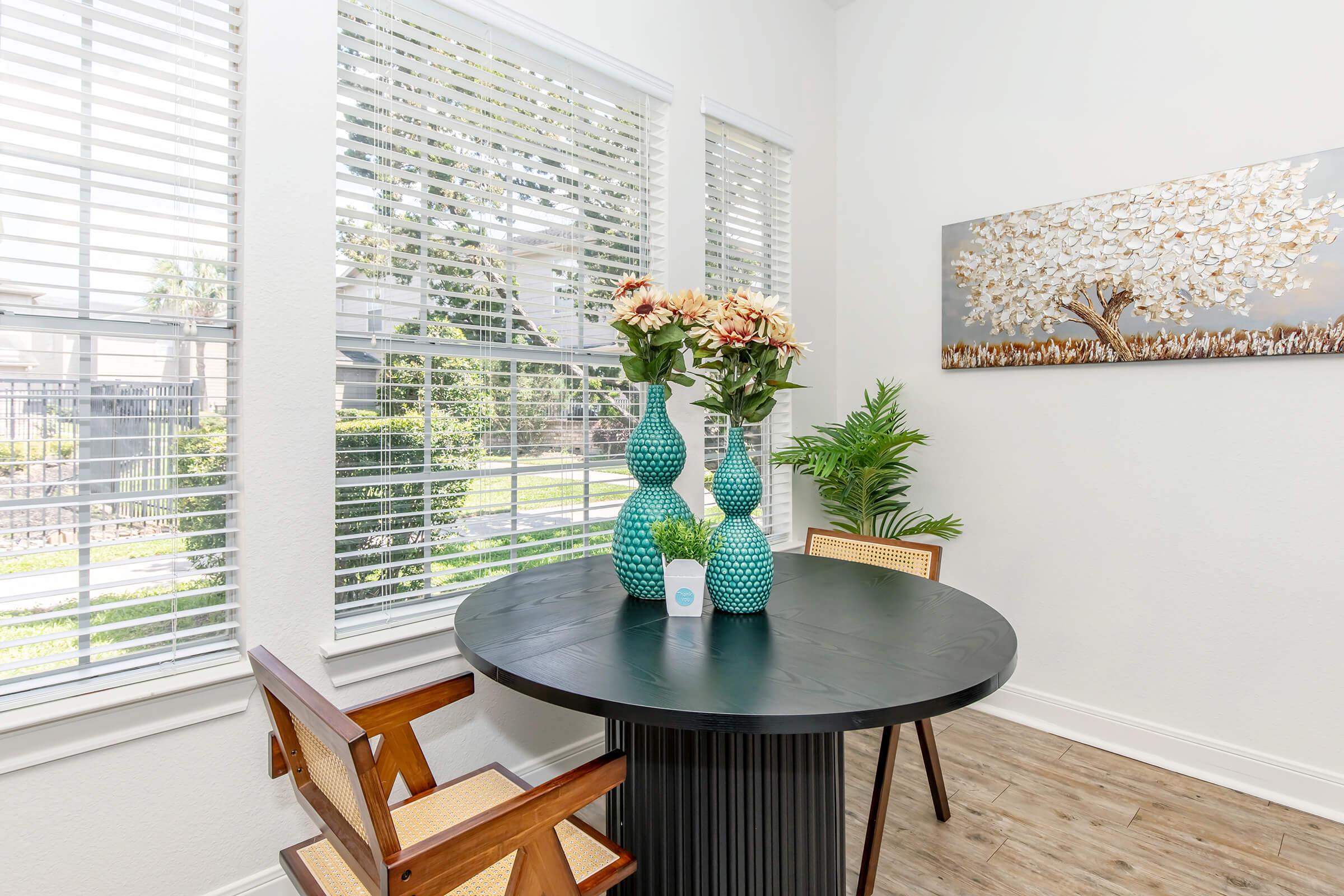 A bright dining area featuring a round black table with two wooden chairs. On the table, there are two blue vases filled with flowers and a small potted plant. The room has large windows with blinds, allowing natural light to illuminate the space, and a wall art piece depicting a tree.