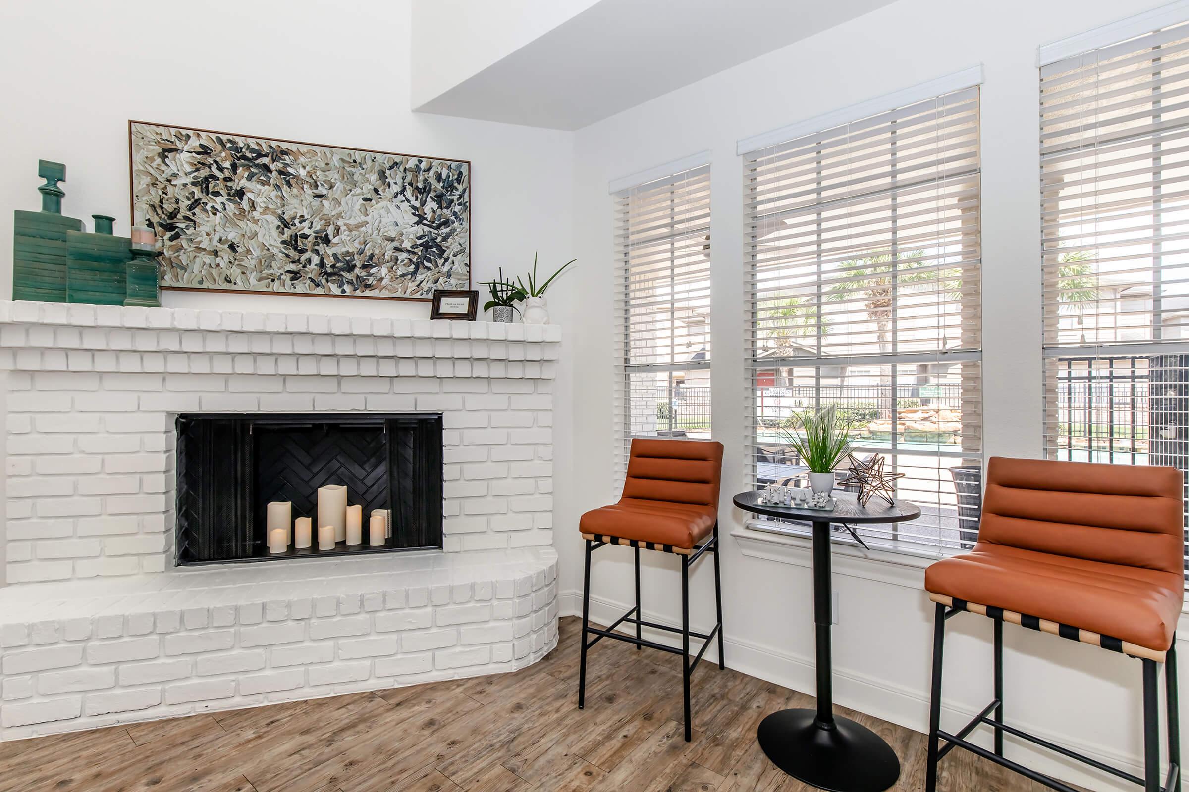 A cozy living space featuring a white brick fireplace with candles, two stylish orange bar stools, and a small round table. Large windows with blinds allow natural light to fill the room, complemented by decorative plants and wall art above the fireplace, creating a warm and inviting atmosphere.