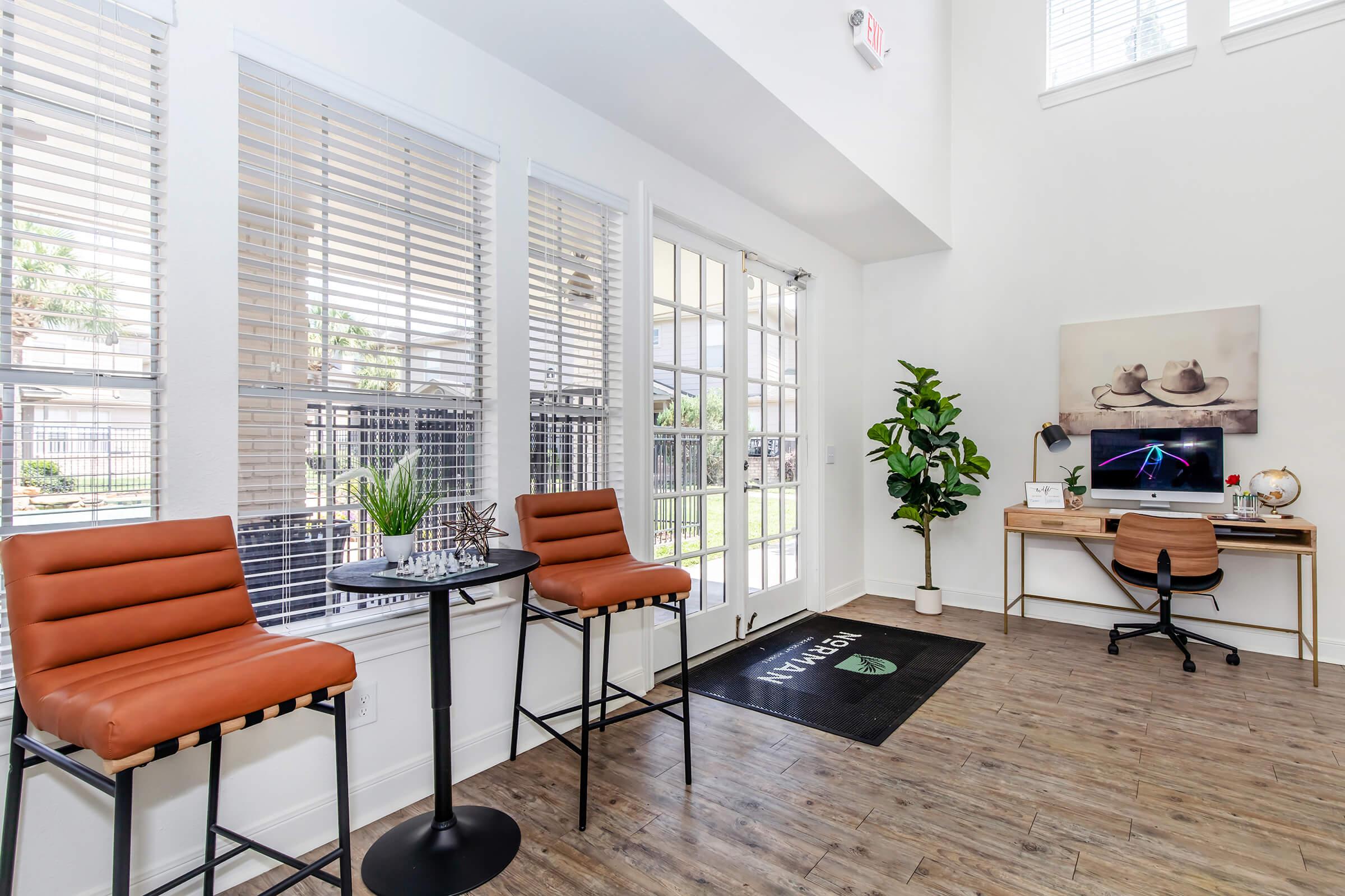 Bright and airy interior space featuring two brown leather barstools at a small table, a green plant in the corner, and a desk with a computer. Large windows with blinds allow natural light to flow in, and there is a welcome mat on the floor. Decorative artwork of hats is displayed on the wall.