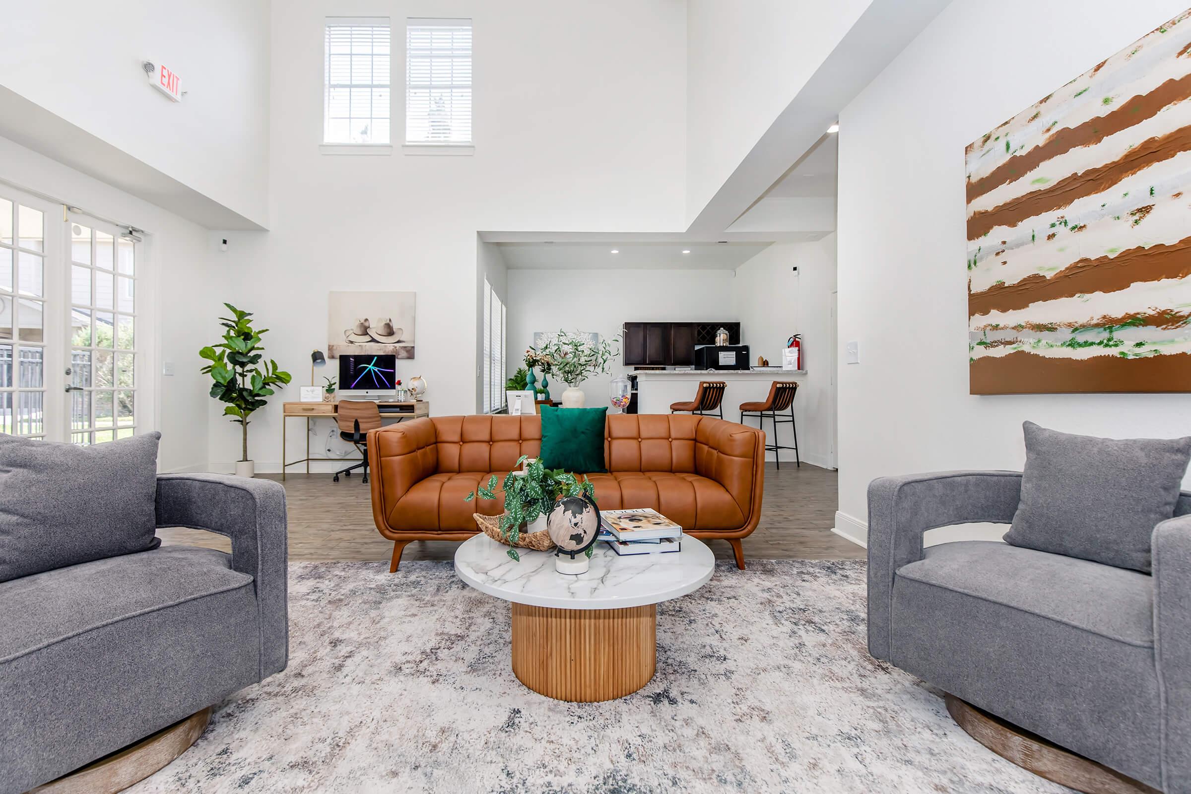 Modern living room with a brown leather sofa and two gray armchairs. A round marble coffee table sits in the center, adorned with decorative items. In the background, there’s a small work desk with a computer, plants, and an open kitchen area, all featuring bright natural light from large windows.