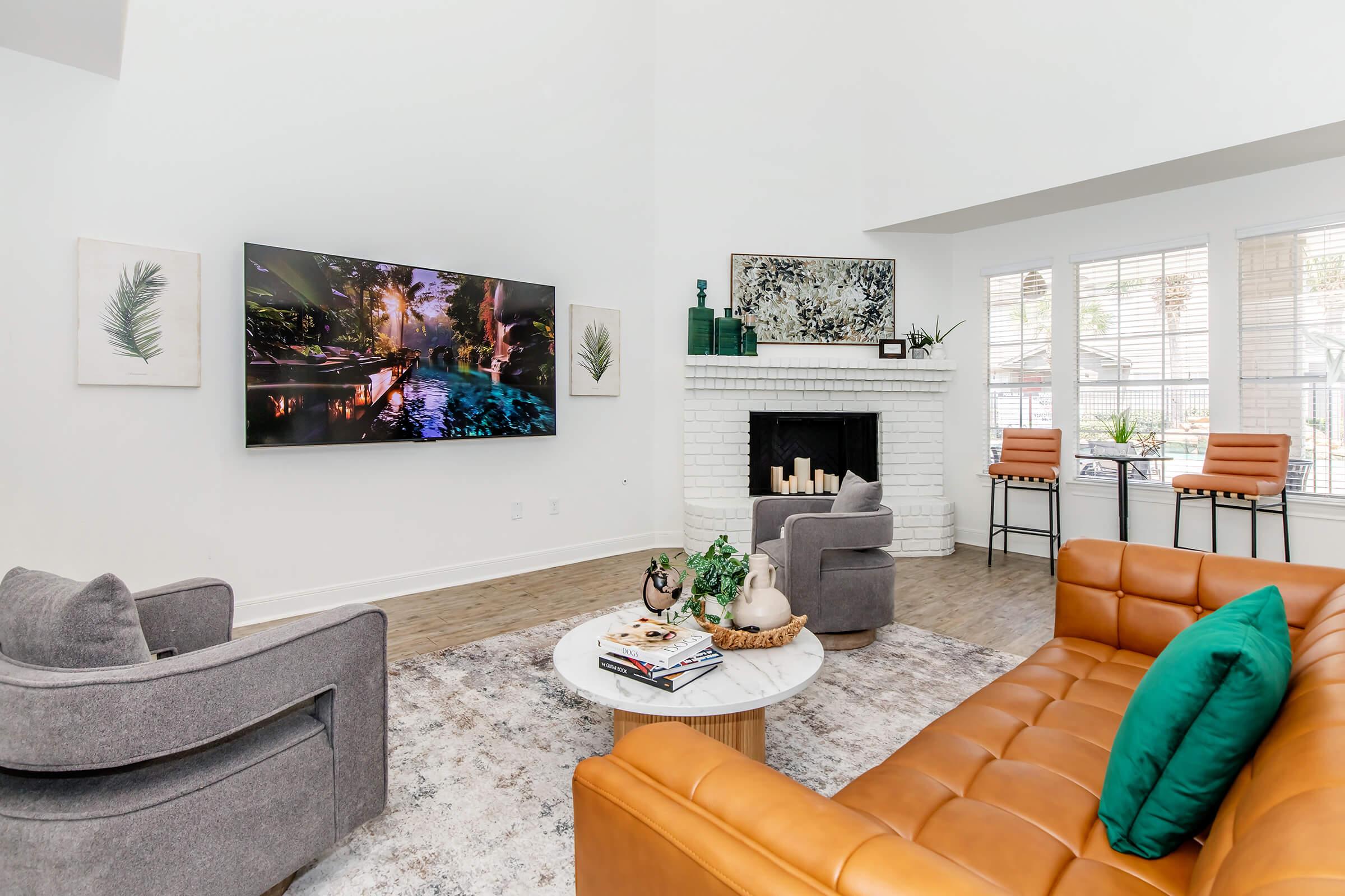 A modern living room featuring a large TV mounted on a white wall, showcasing a vibrant nature scene. It includes a brown leather sectional sofa with a green pillow, two grey armchairs, a round coffee table with decorative items, and a fireplace with candles. Large windows provide natural light.