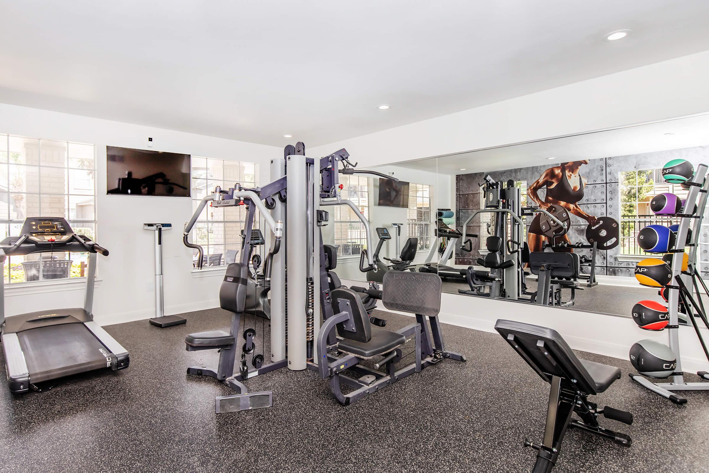 A modern gym interior featuring a variety of exercise equipment including a multi-station weight machine, a treadmill, and free weights. Large mirrors line the wall, and a wall mural depicting a muscular athlete adds a motivational touch to the space. Bright, natural light filters through the windows.