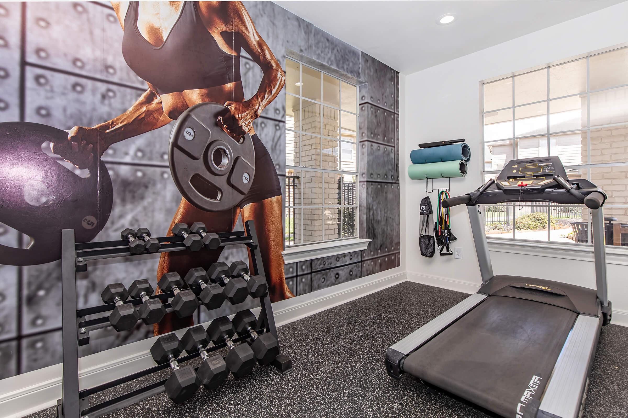 A fitness room featuring a treadmill and a rack of dumbbells. The wall is adorned with a large mural of a muscular person lifting weights. There are also yoga mats and exercise accessories on a nearby wall, creating an inspiring workout environment. Natural light enters through two windows.